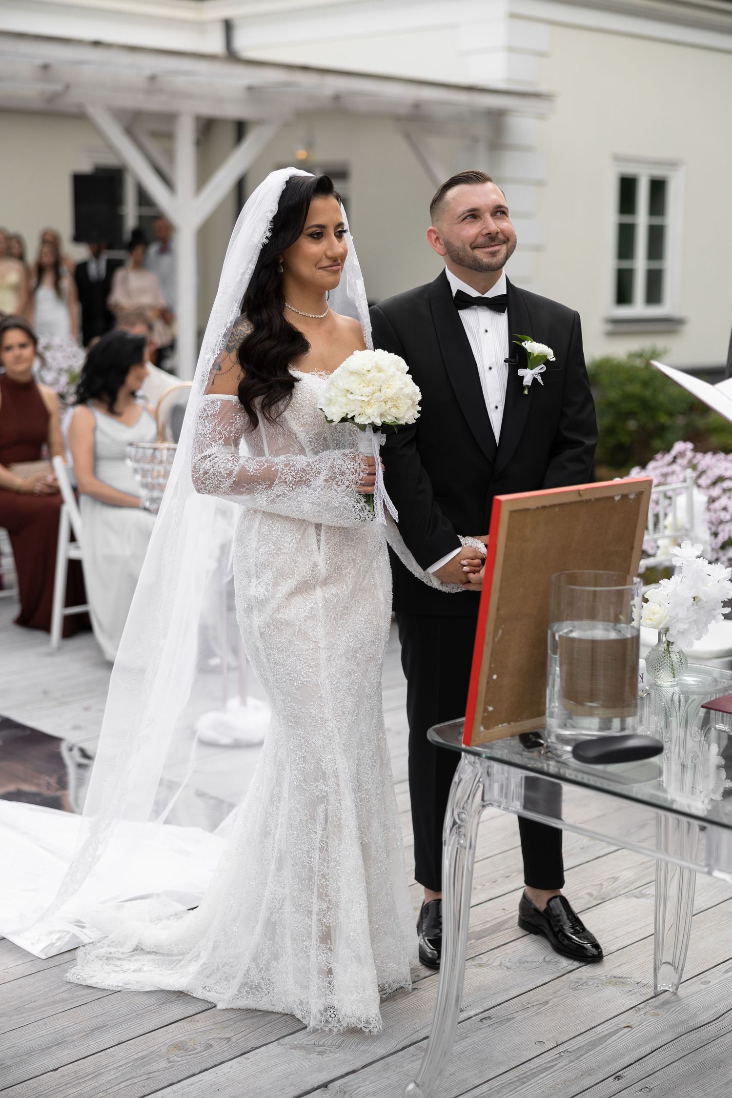 Bride in white lace gown holding bouquet stands with groom in black tuxedo during outdoor wedding ceremony.
