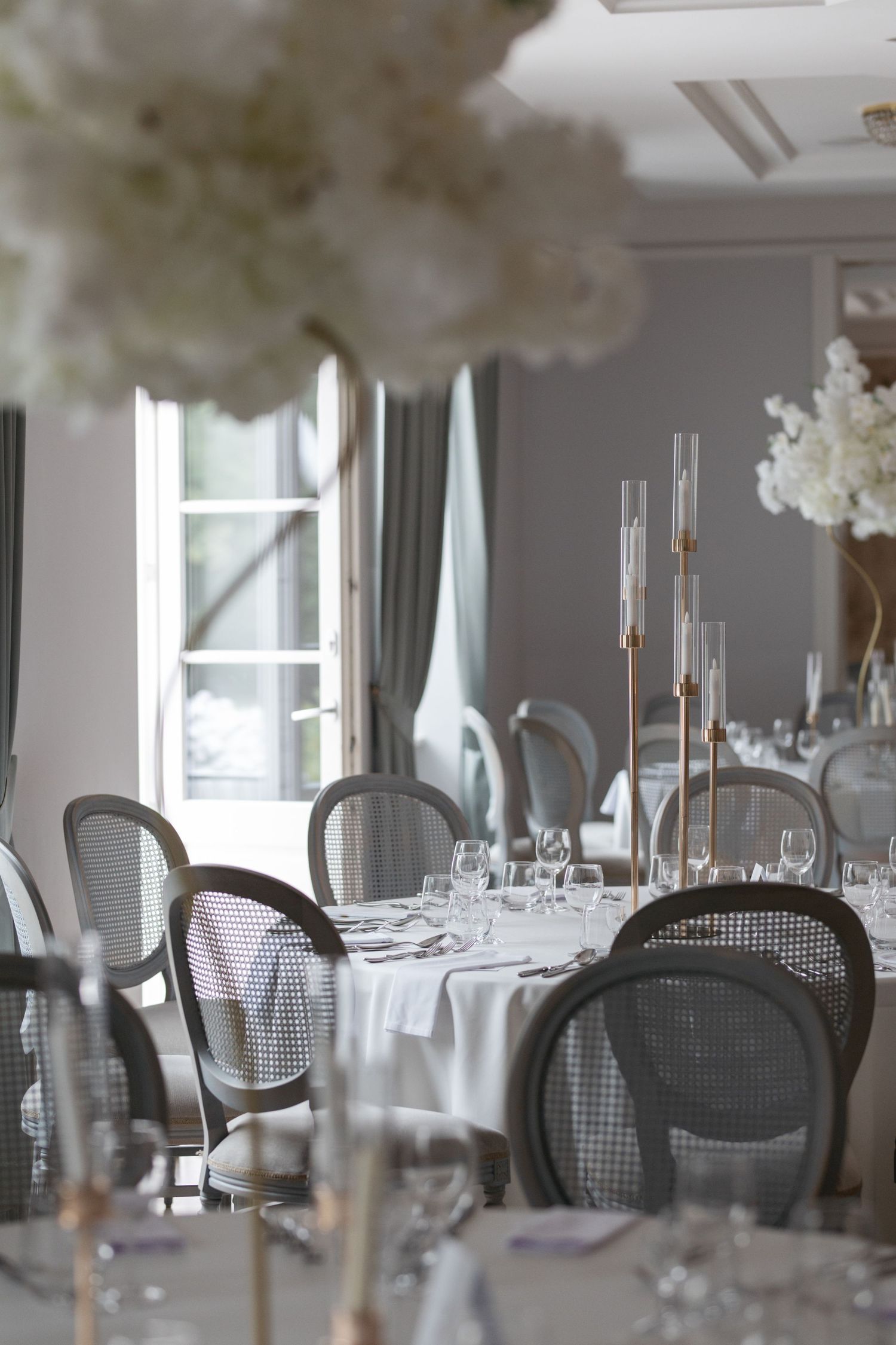 Elegant wedding reception venue with white floral centerpieces, tall gold candelabras, and classic cane-back chairs around draped tables.