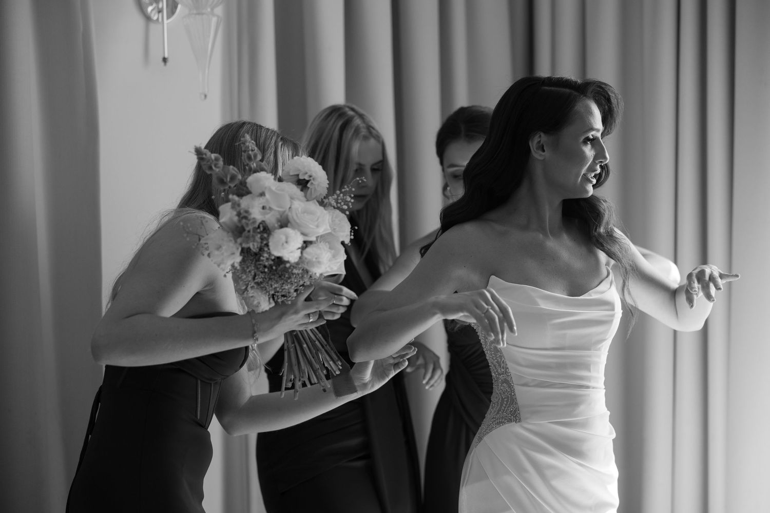 Black and white photo of a bride in white gown with bridesmaids helping her prepare, holding a floral bouquet.