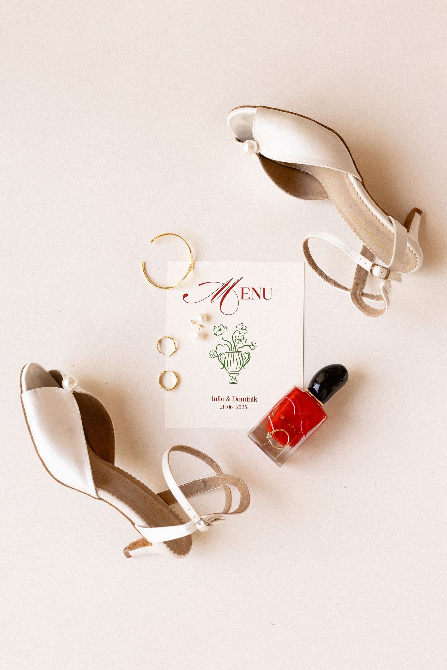 Wedding day essentials featuring elegant white heels, gold wedding rings, a red nail polish bottle and delicate menu card on beige surface.