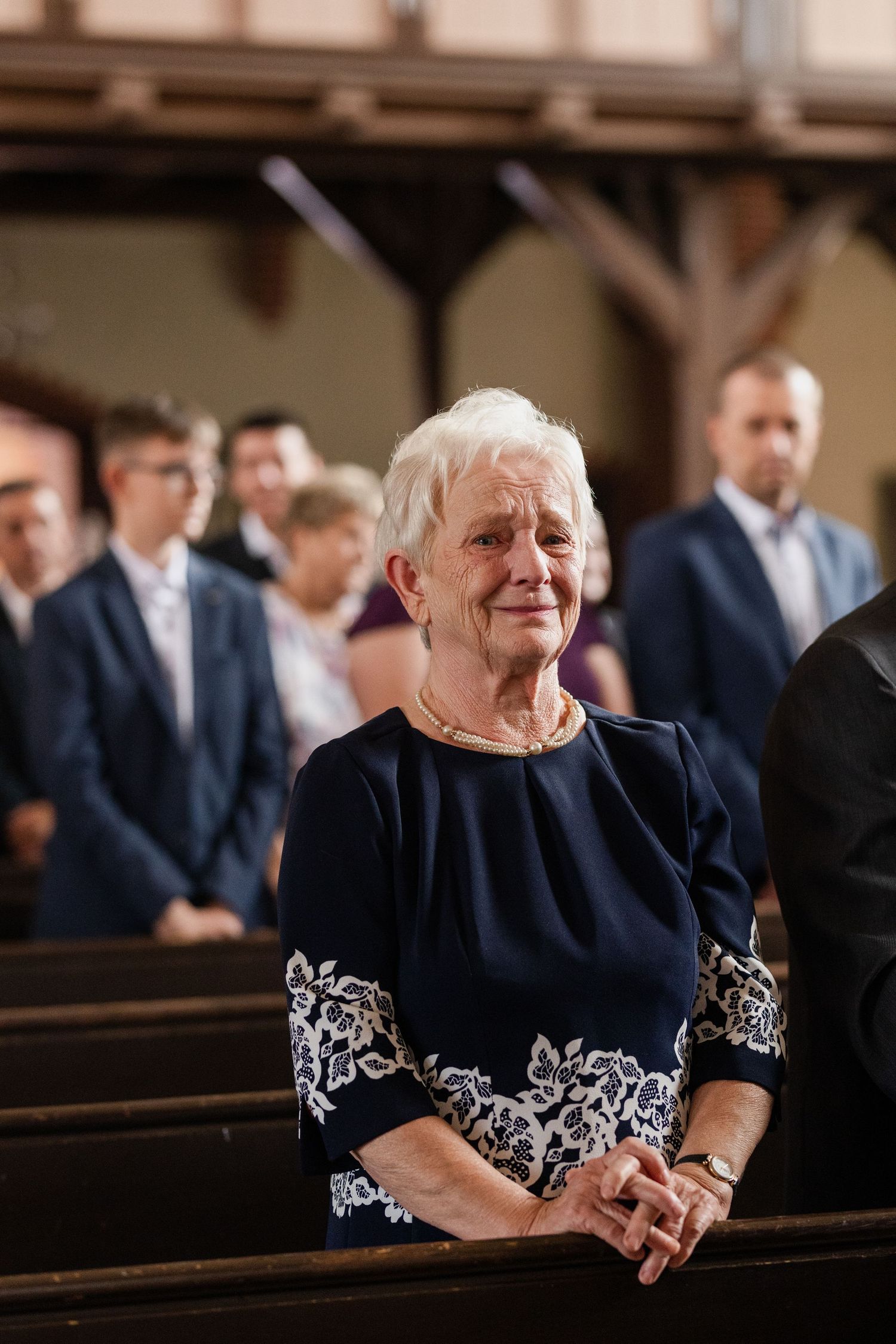 An elderly woman in a navy dress with white embroidery laughs joyfully while seated in a church pew.