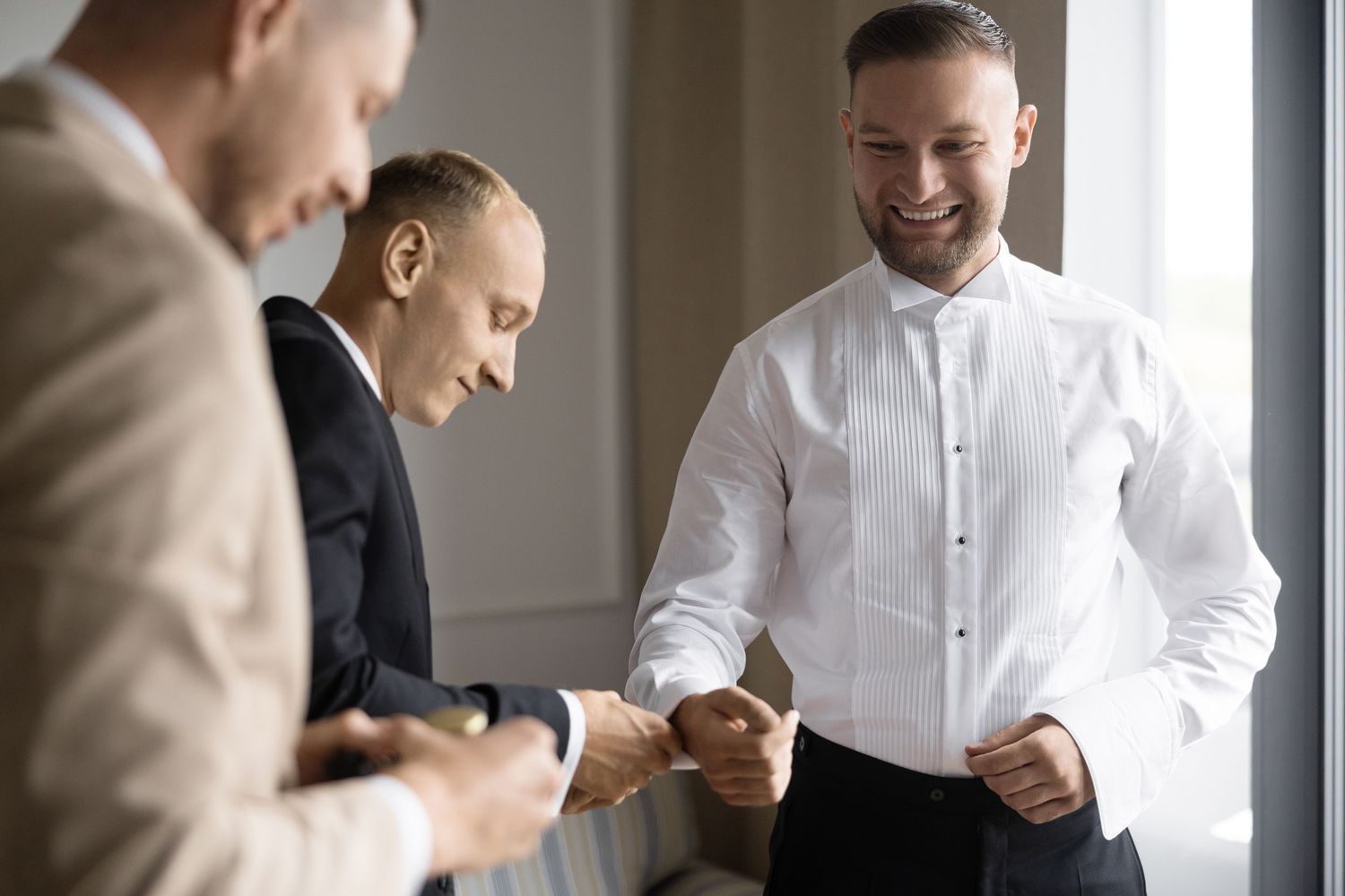 Three men in formal attire smiling and shaking hands, preparing for a wedding celebration indoors.
