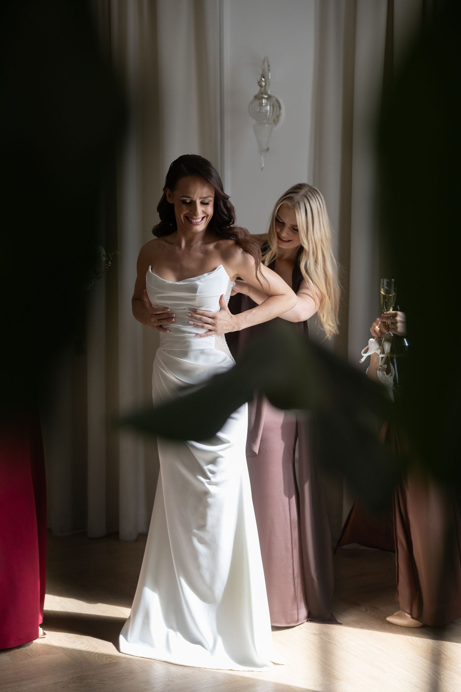 A bride in a white satin gown smiles as a blonde bridesmaid in mauve helps her dress before the wedding ceremony.