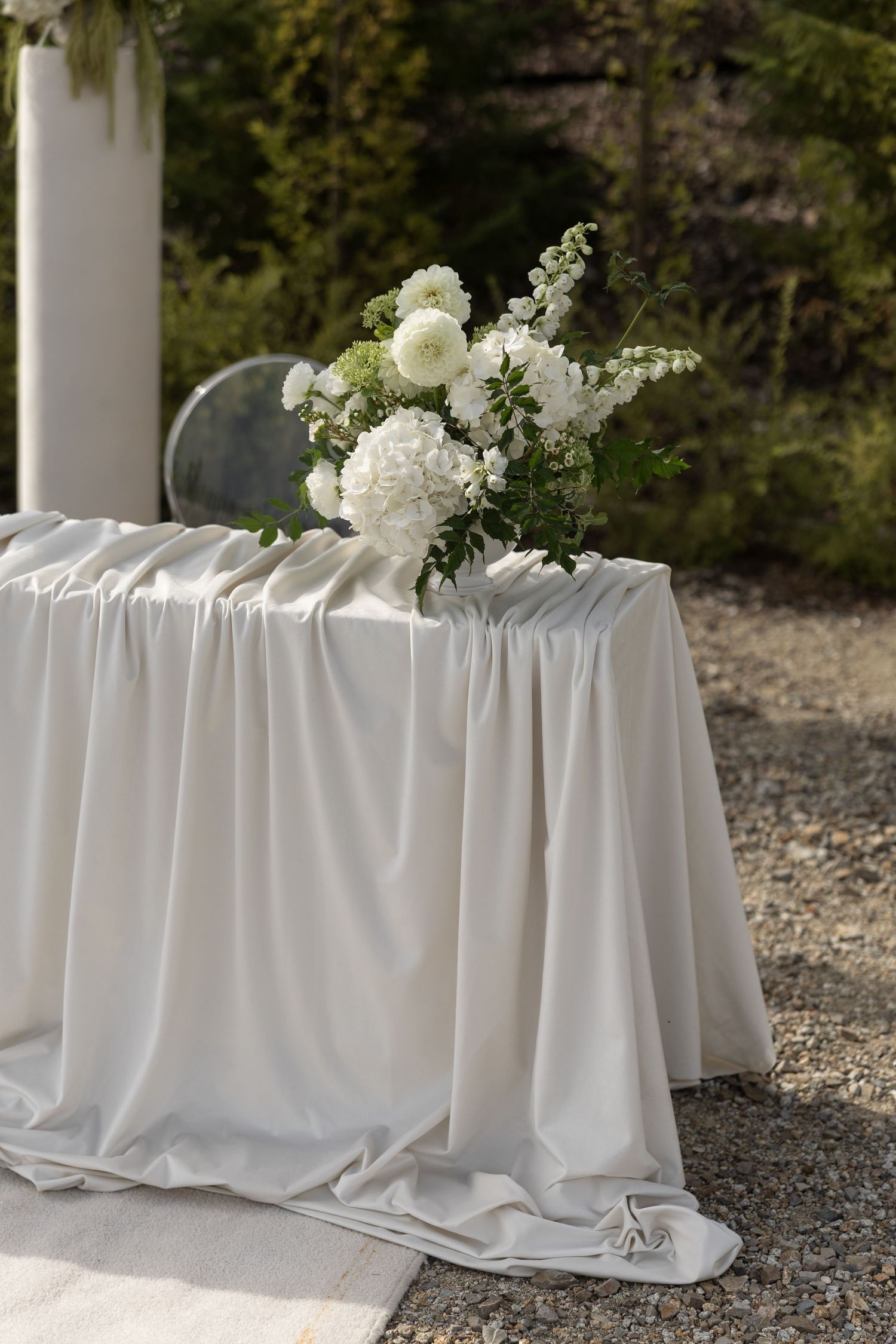 Elegant outdoor wedding table with white draped linen and white floral arrangement of dahlias and greenery.