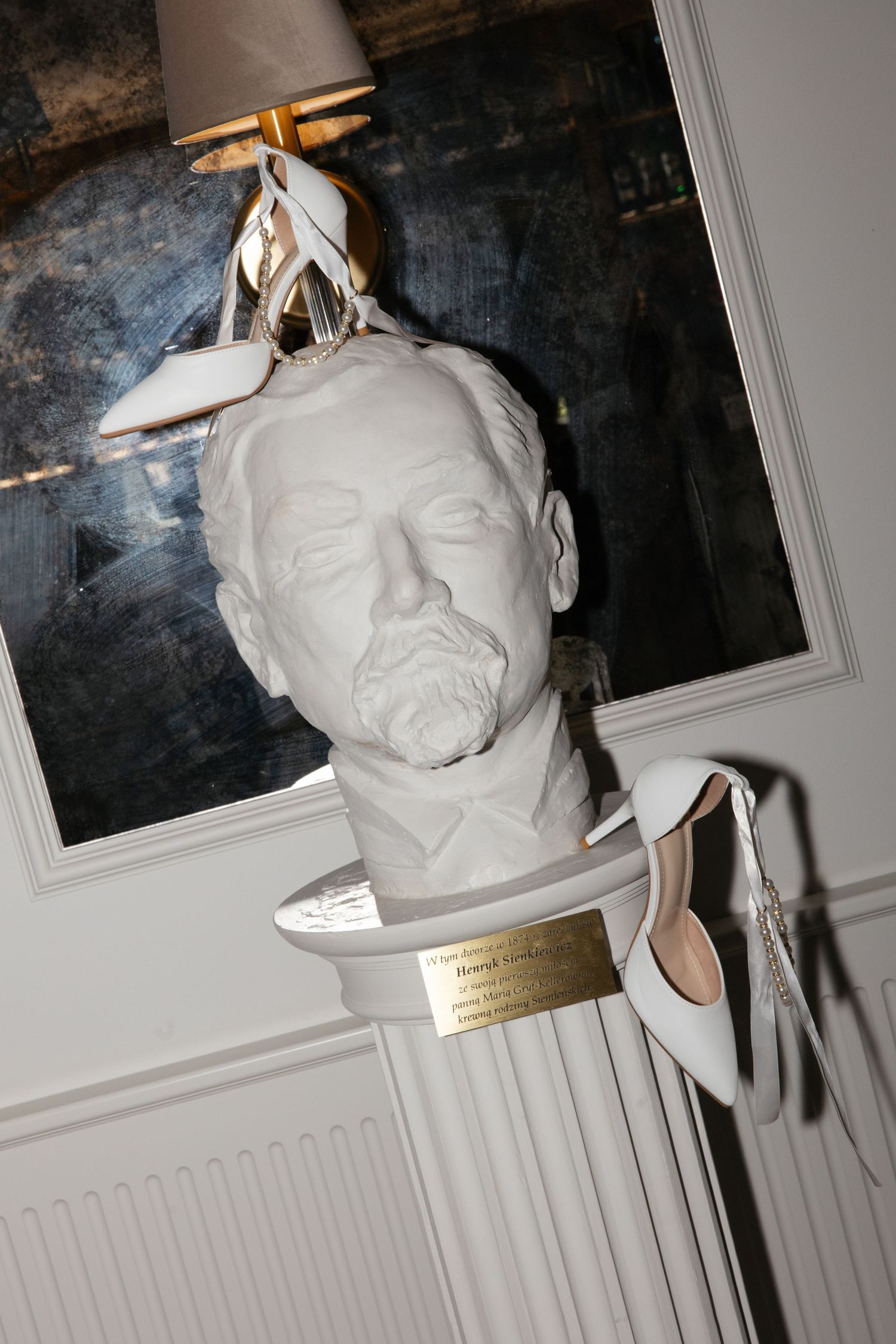 White plaster bust sculpture wearing white high heel shoes as a hat, displayed on a gold placard pedestal.