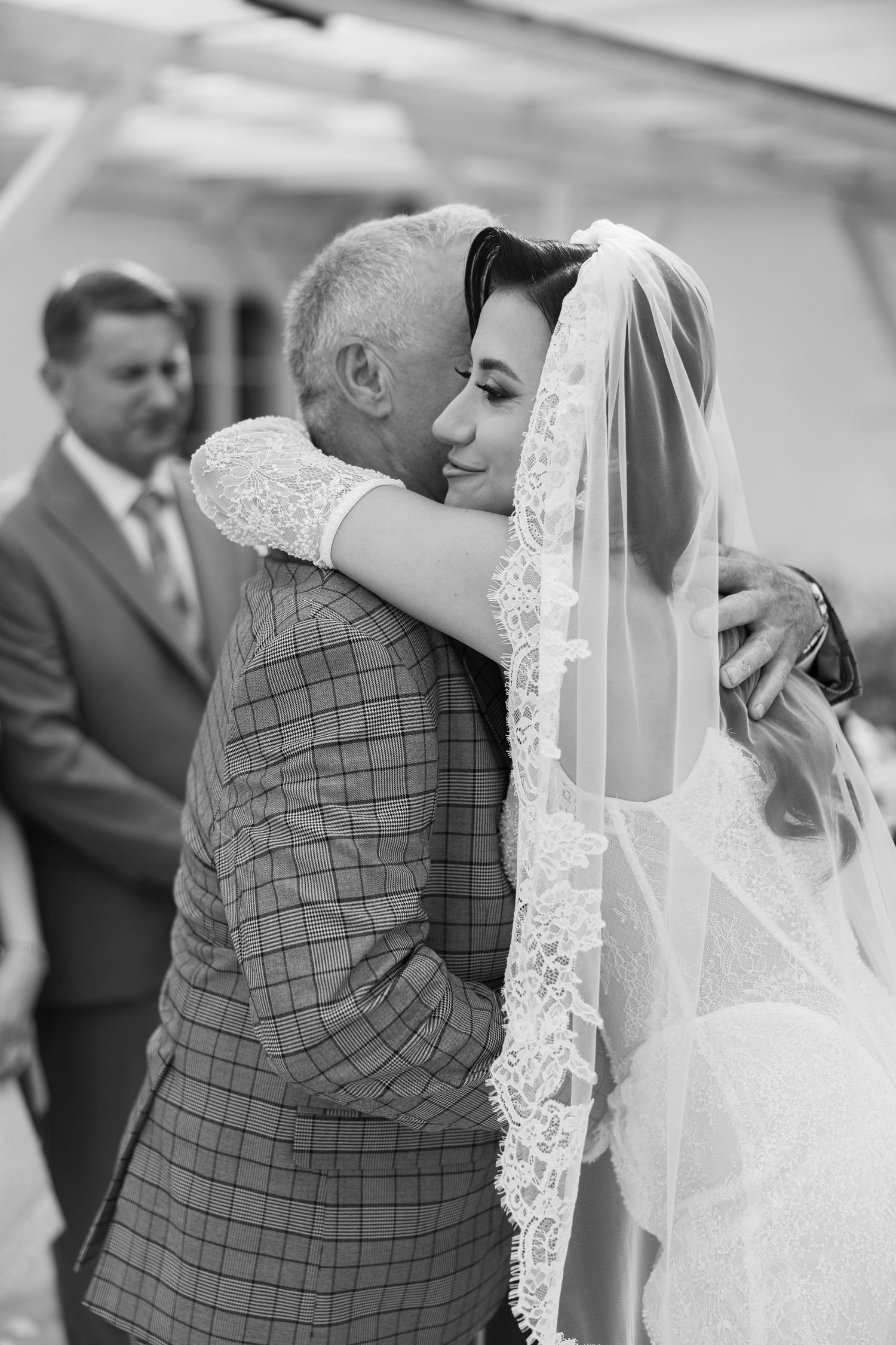 Black and white photo of a bride in lace veil embracing an older man in a plaid suit at a wedding ceremony.