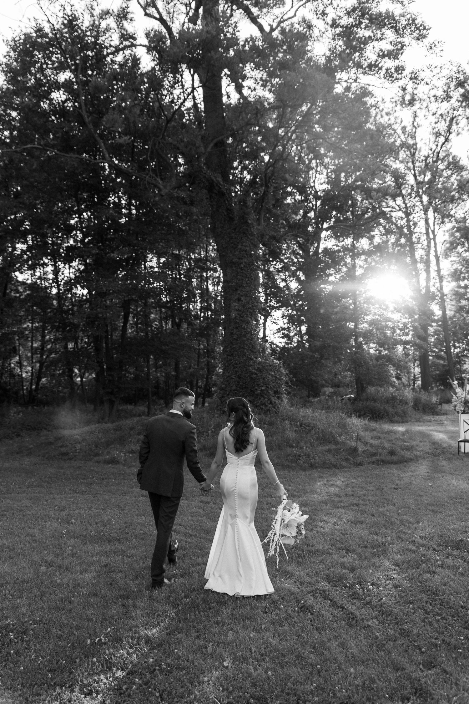 A couple in wedding attire walks together through a sunlit meadow surrounded by trees in a romantic black and white photo series.