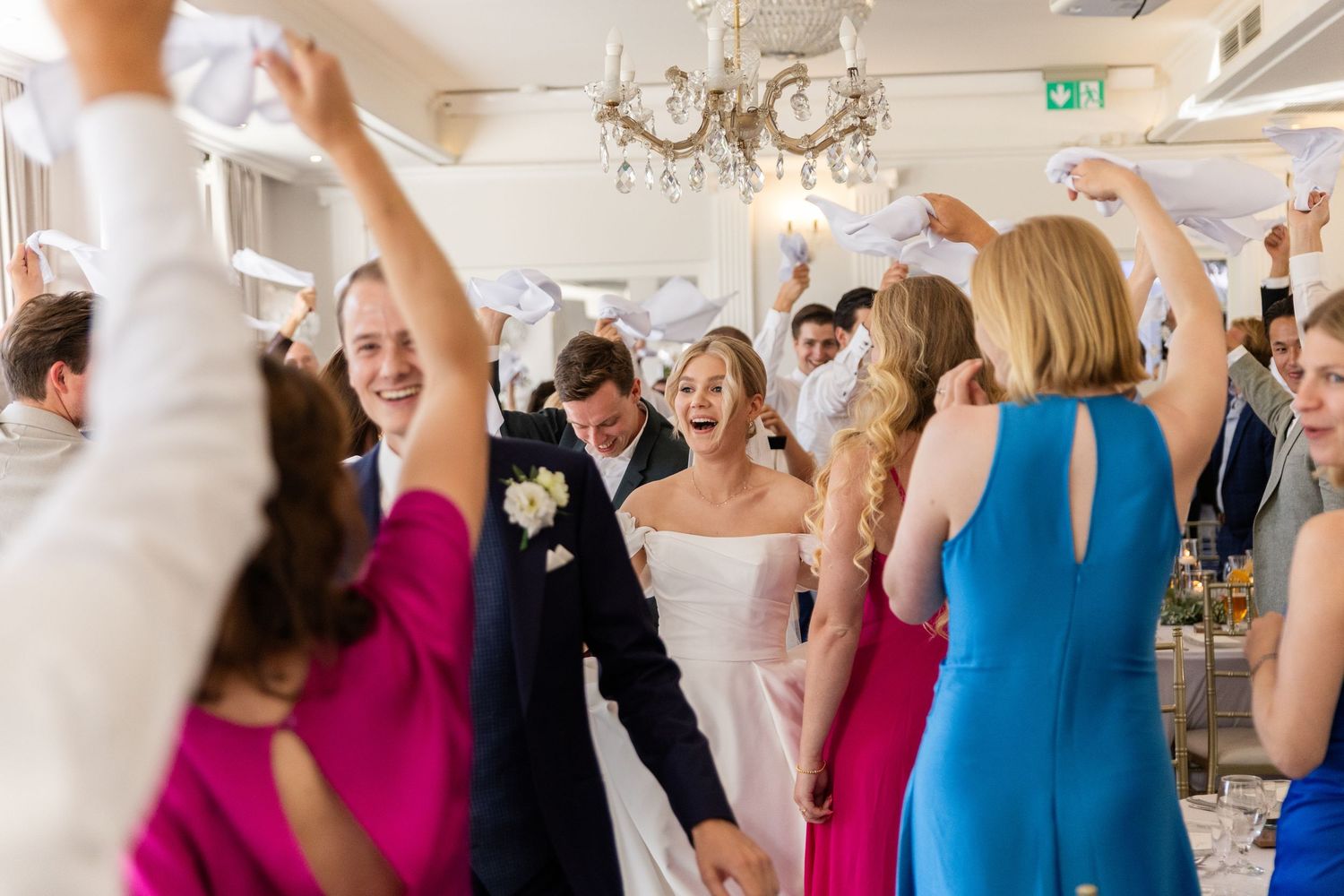Wedding party guests dancing and celebrating joyfully in an elegant white reception venue with chandelier.