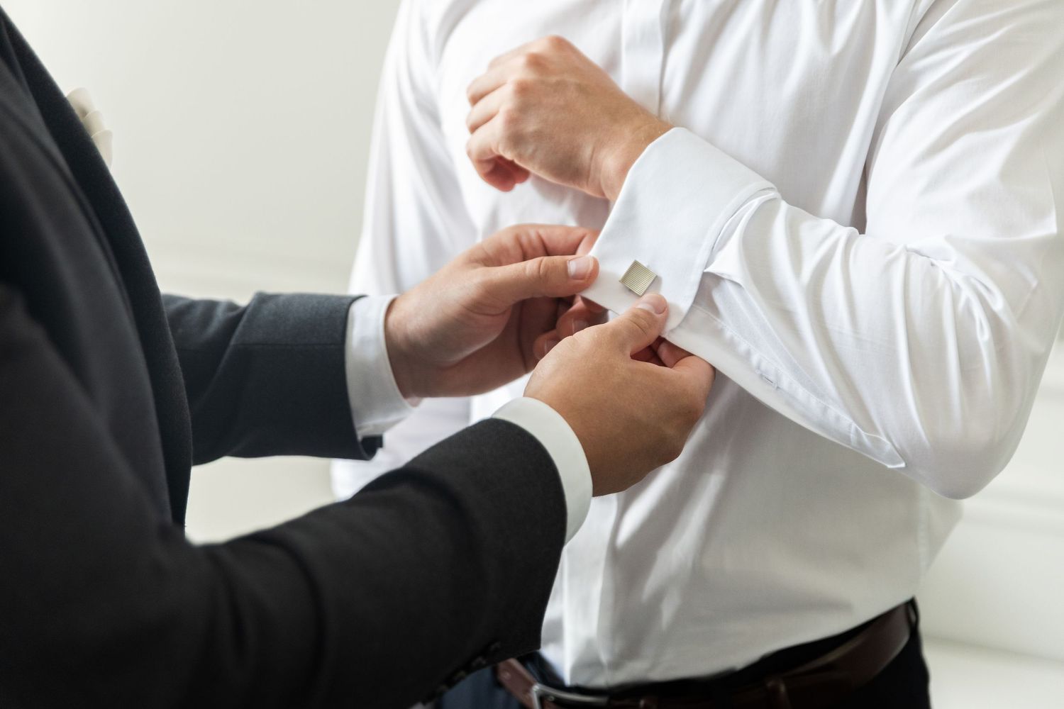Close-up of hands adjusting a white dress shirt sleeve and cufflink against a light background.