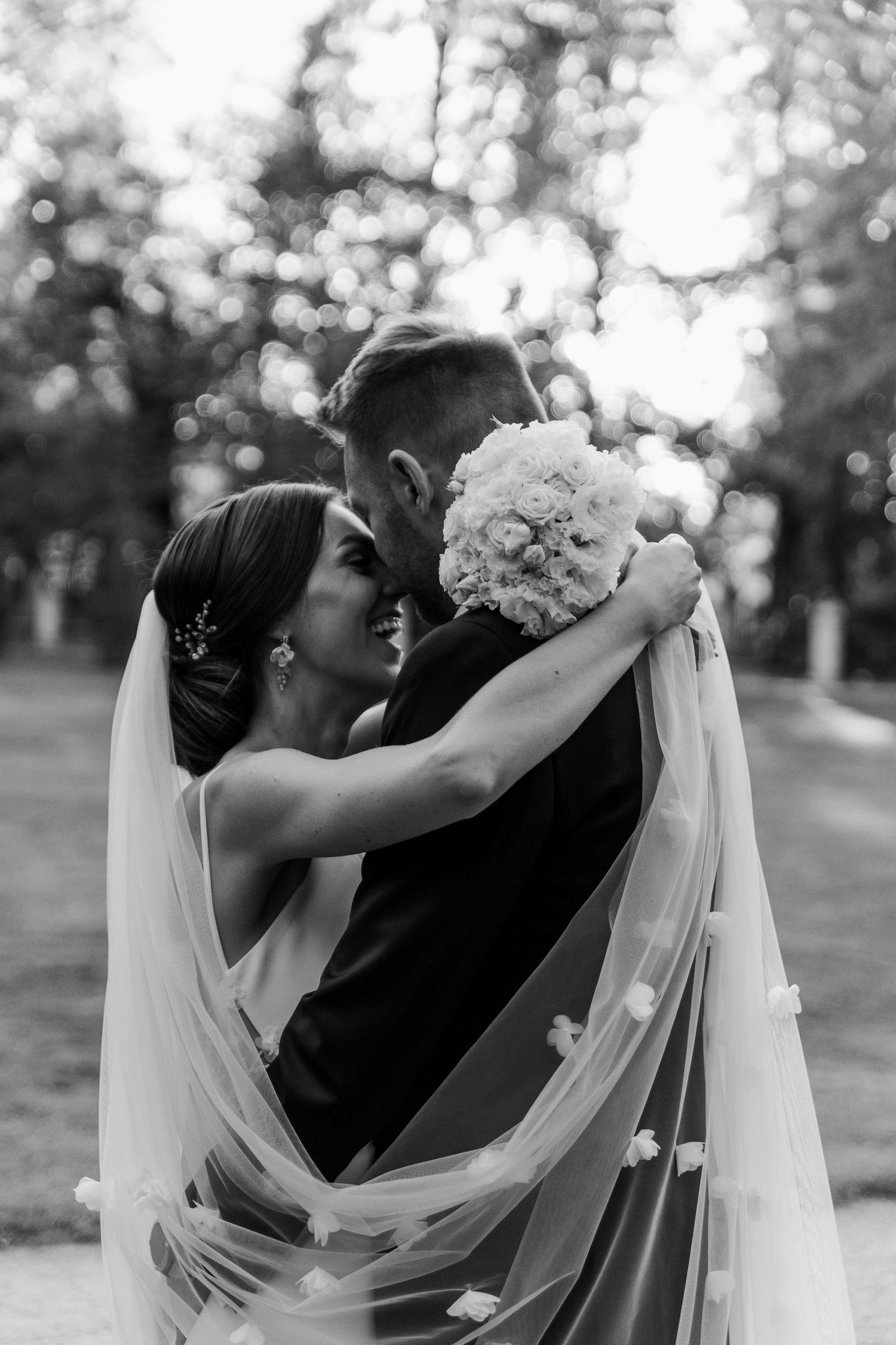 Black and white wedding photo showing couple embracing with flowing veil and white bouquet.