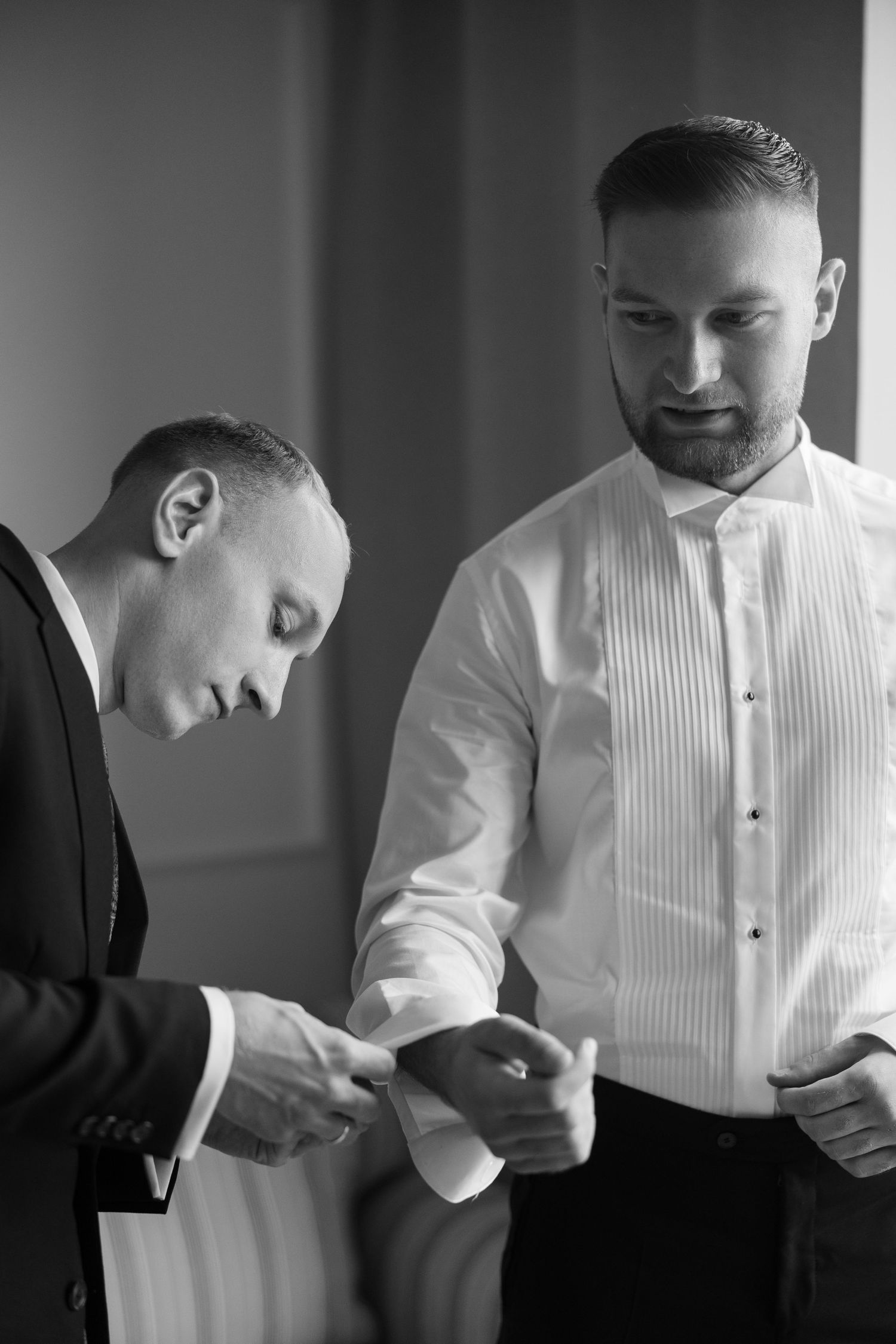 Black and white photo of two men in formal attire, one helping the other with cufflinks before a wedding ceremony.