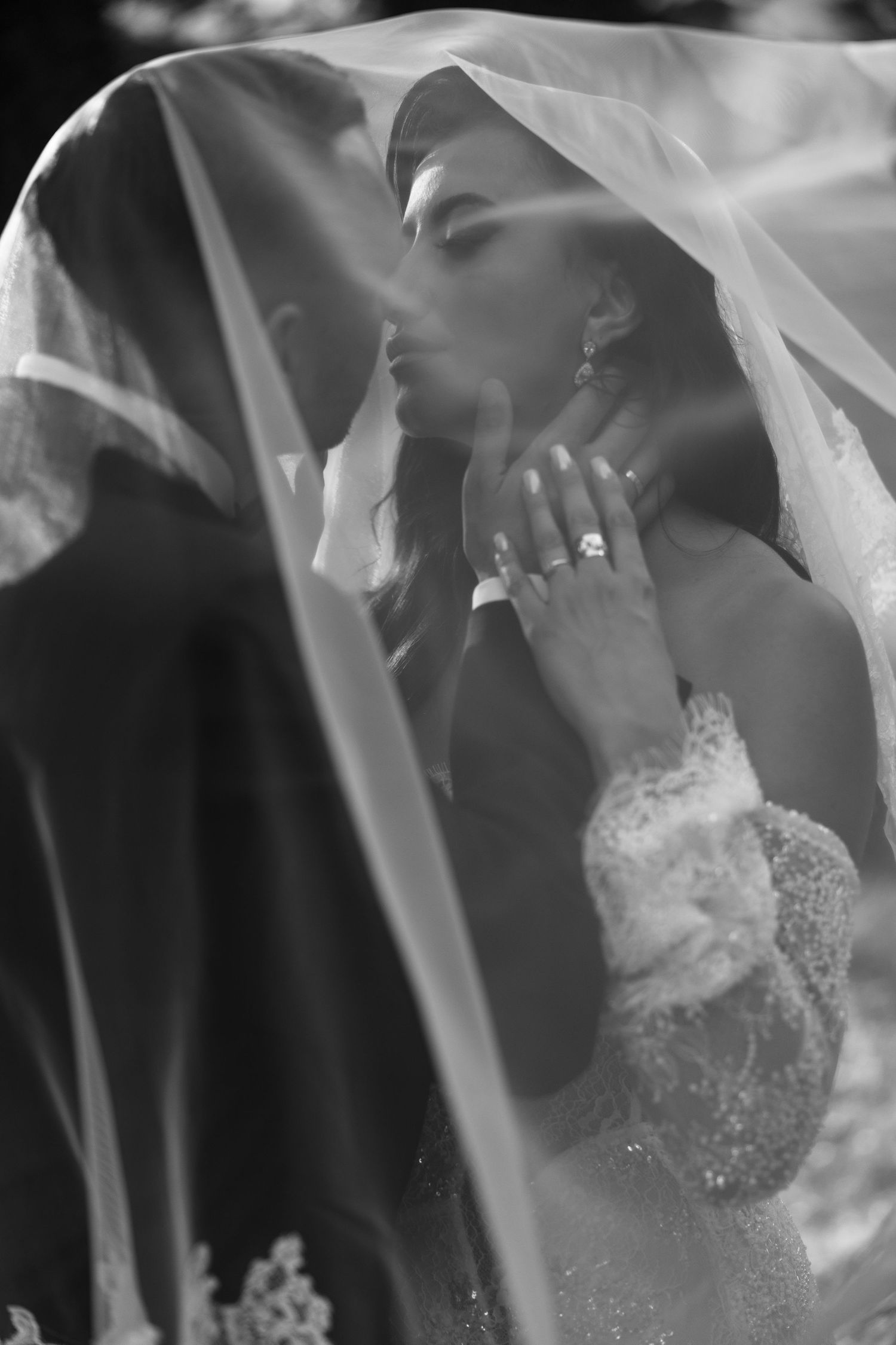 Black and white wedding photo of bride and groom kissing under a veil, bride wearing lace dress and diamond ring.