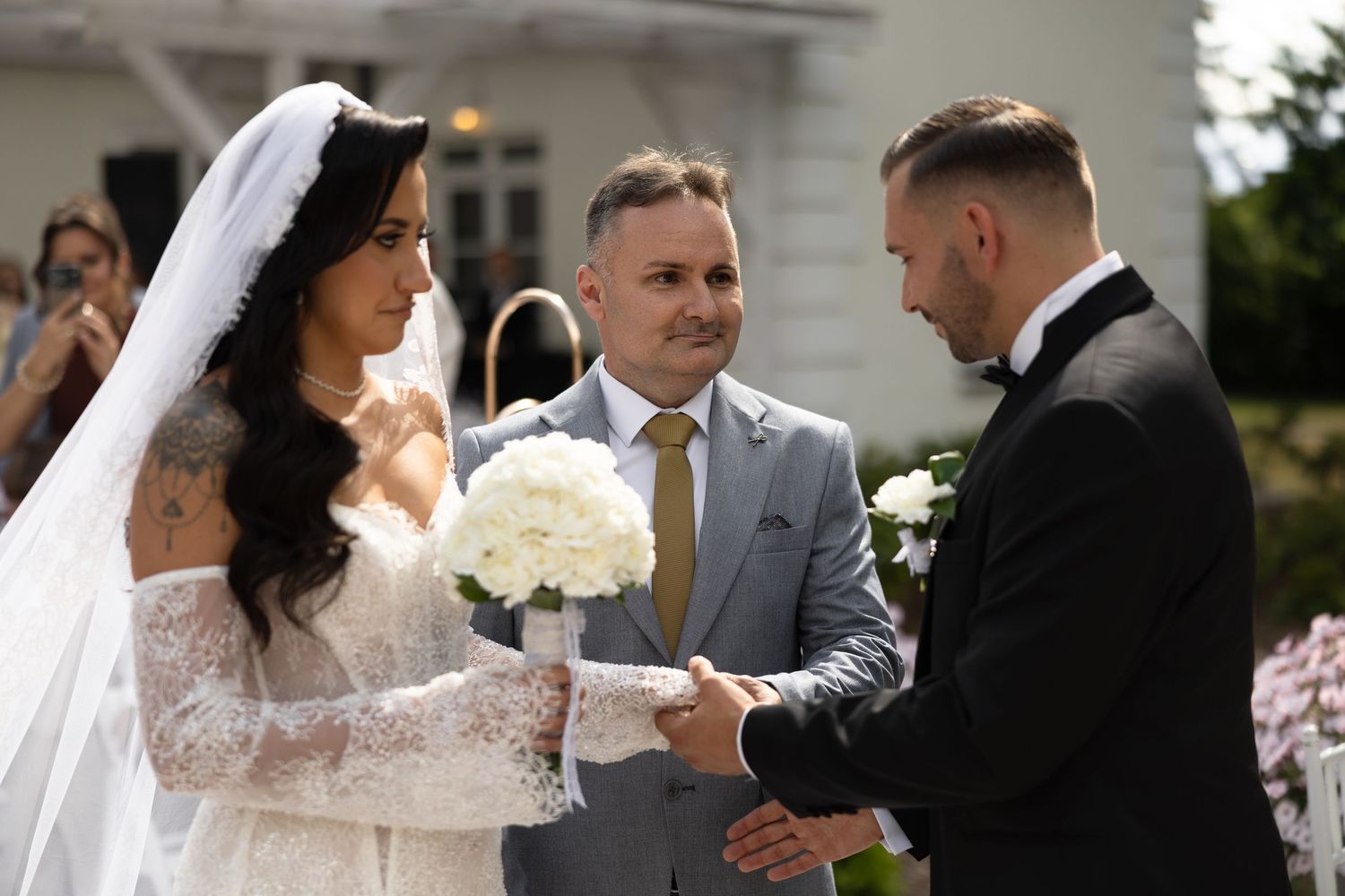 Bride and groom exchange vows outdoors with officiant in gray suit during elegant wedding ceremony.