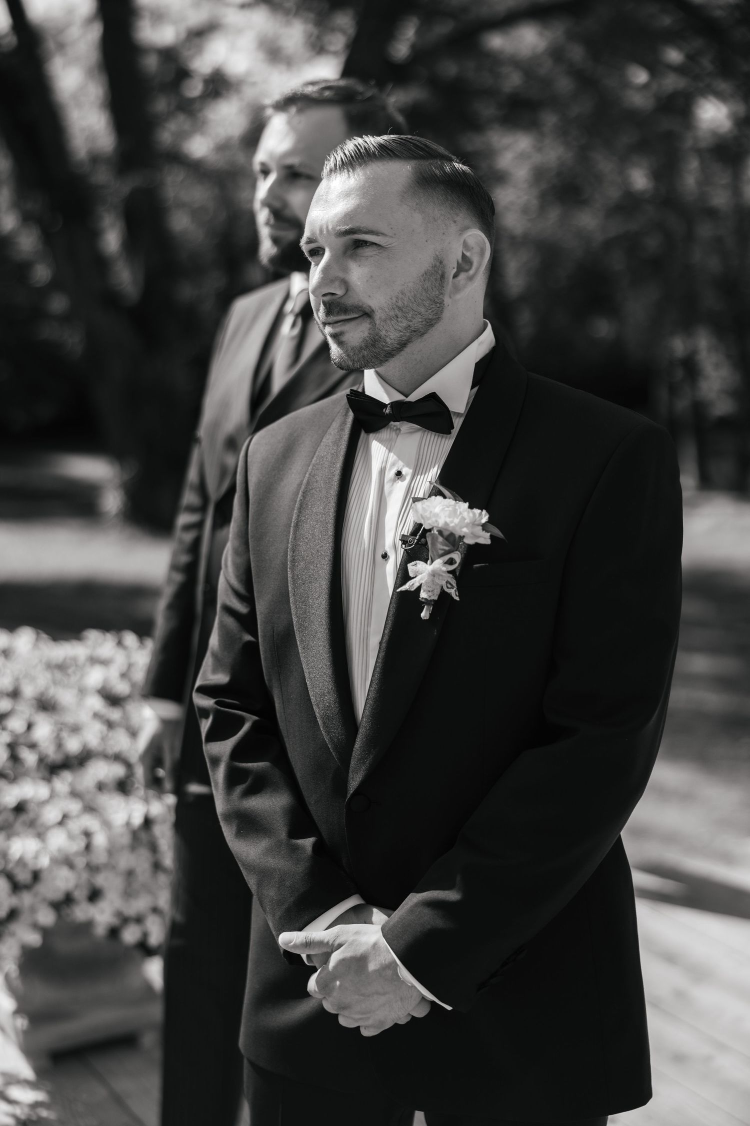 Black and white photo of a groom in a tuxedo with boutonniere standing at the altar, another man visible behind him.