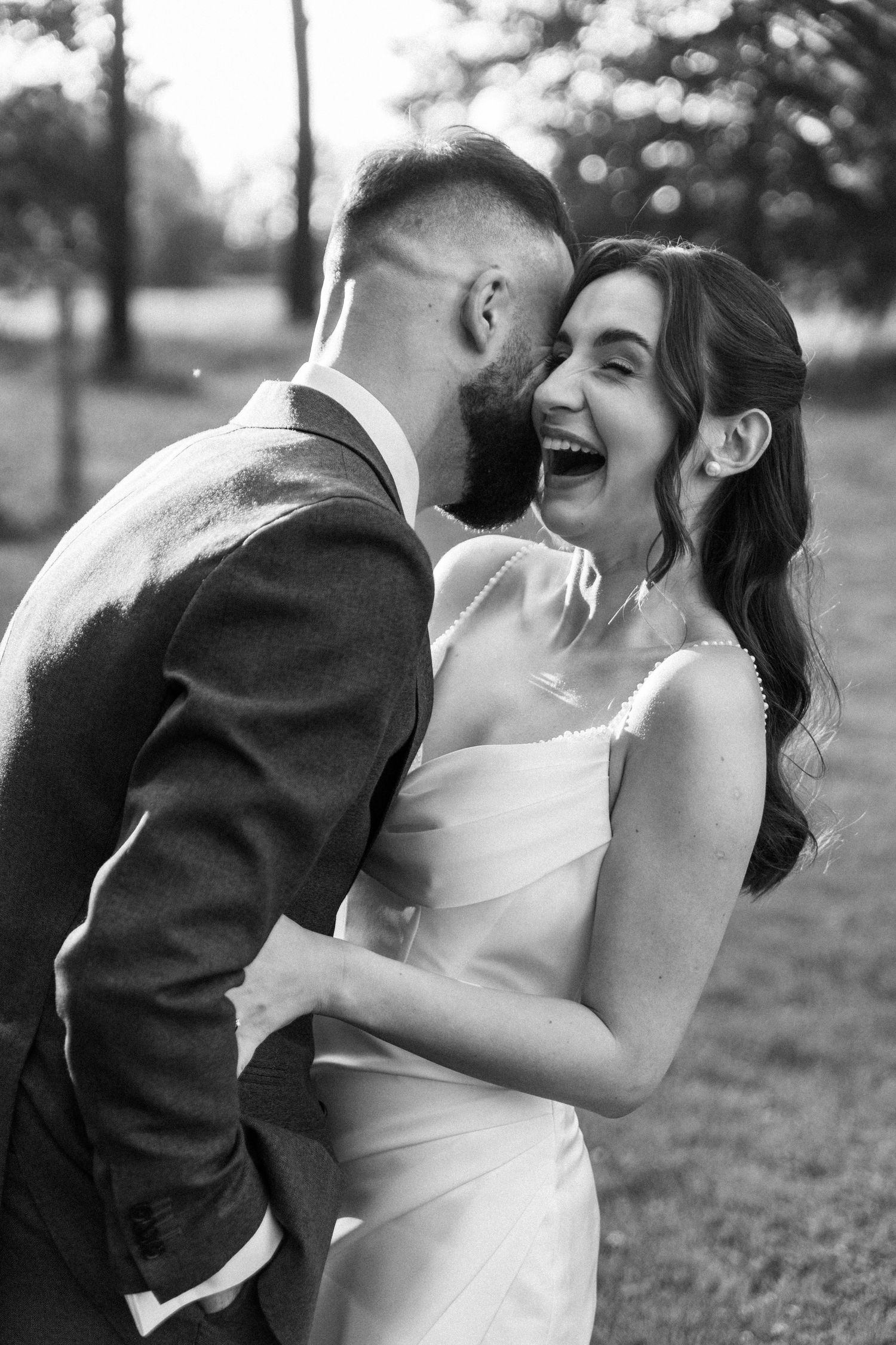 Black and white photo of a joyful wedding moment outdoors with bride laughing in elegant white dress.