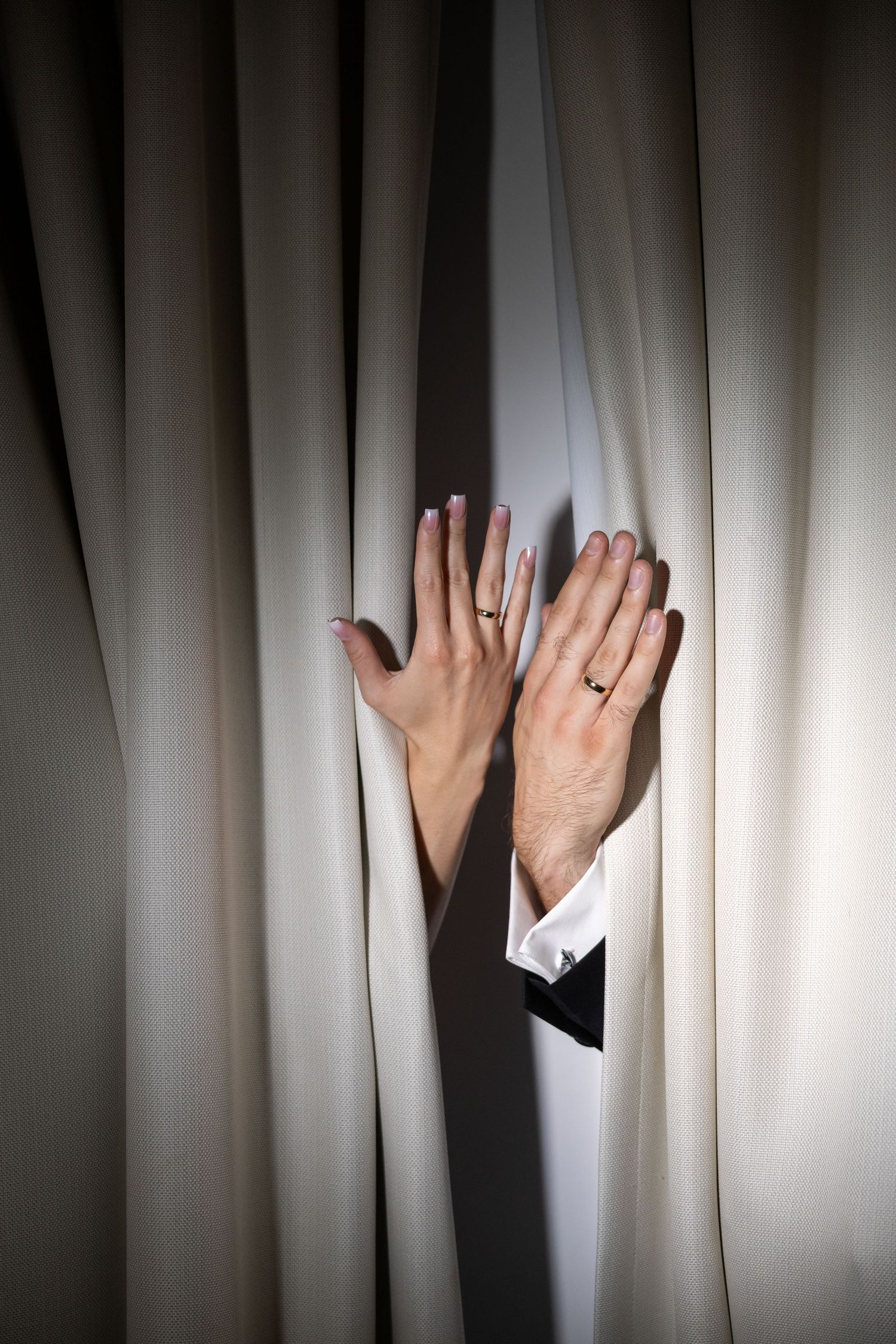 Two hands with wedding rings emerge through cream curtains, symbolizing a couple's union.