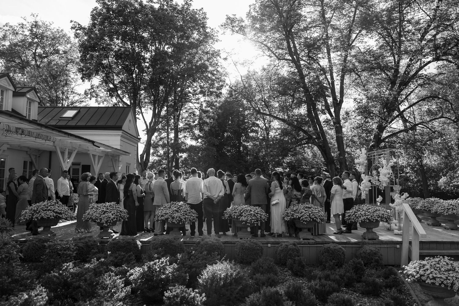 Black and white photo of guests gathered outdoors at an elegant garden wedding reception surrounded by trees and flowers.