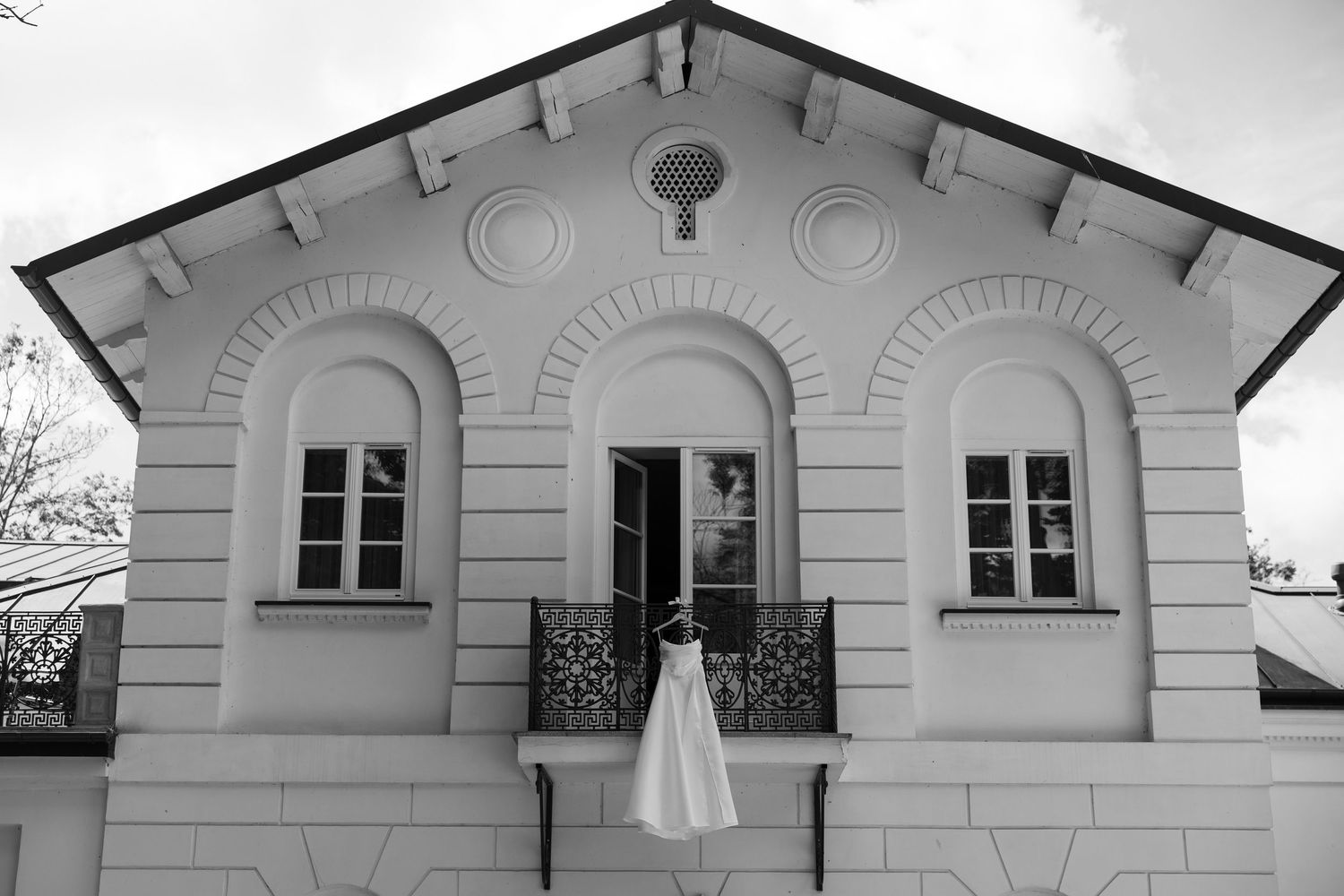 Black and white photo of a wedding dress hanging on ornate balcony of elegant white classical building.