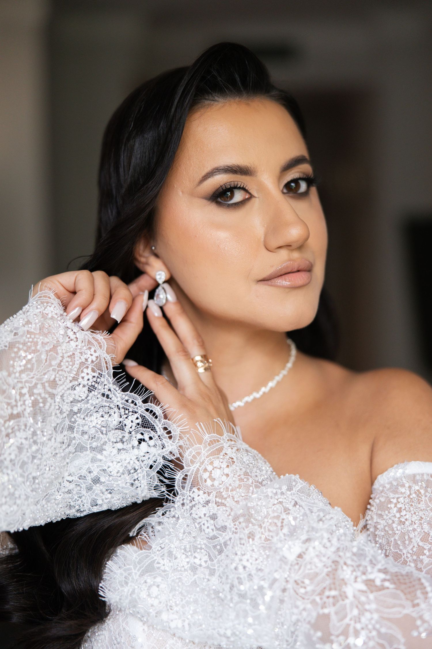 Bride in white lace gown adjusting pearl earring, wearing pearl necklace and gold rings, with elegant bridal makeup.