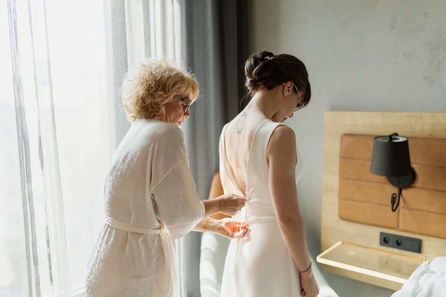 Two women stand by a window while helping with dress preparations for a formal event.