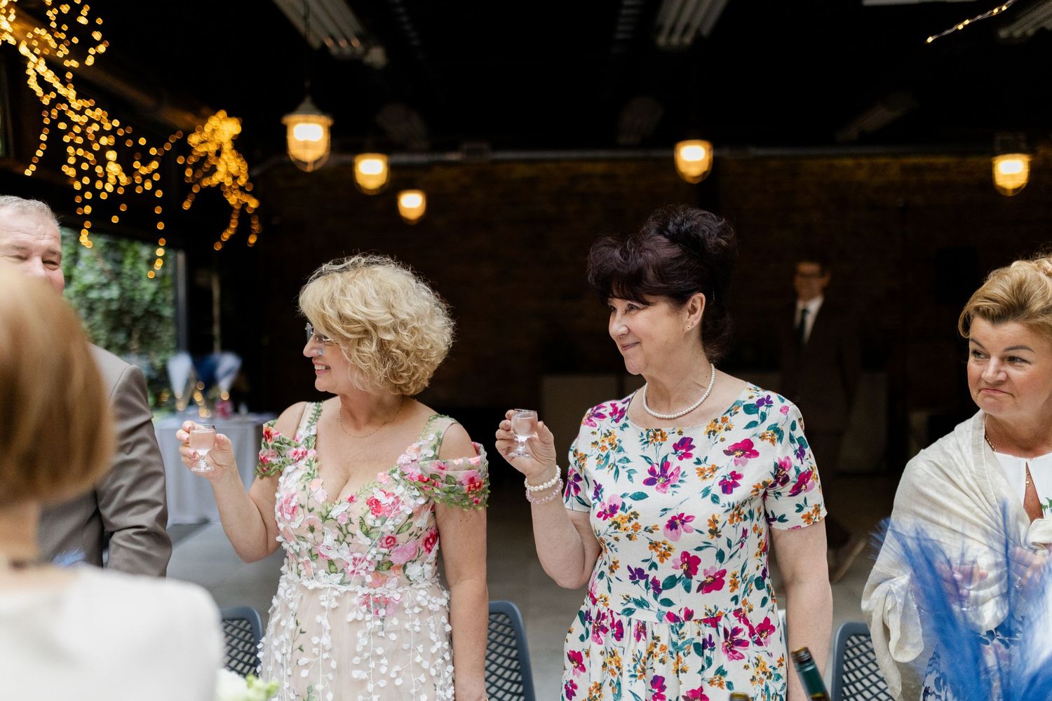 Guests in floral dresses dance and socialize at an evening reception with twinkle lights and lanterns overhead.