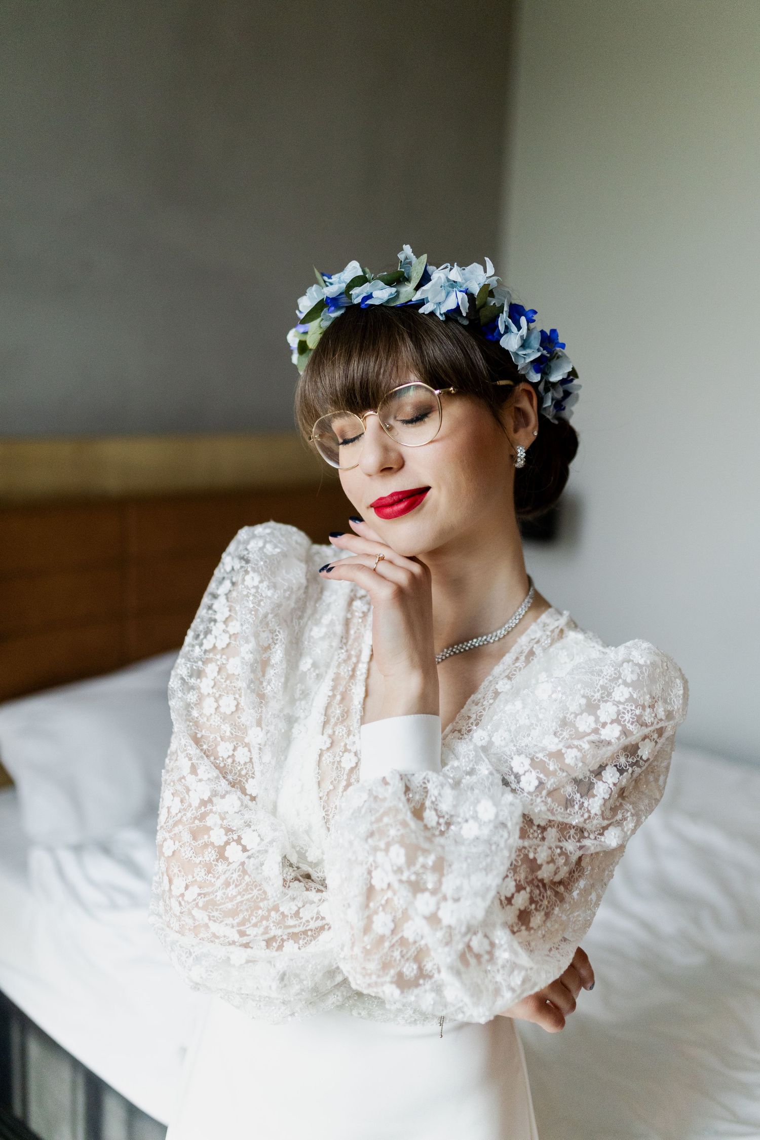 Elegant bridal portrait featuring white lace dress with long sleeves and blue floral crown against light interior.