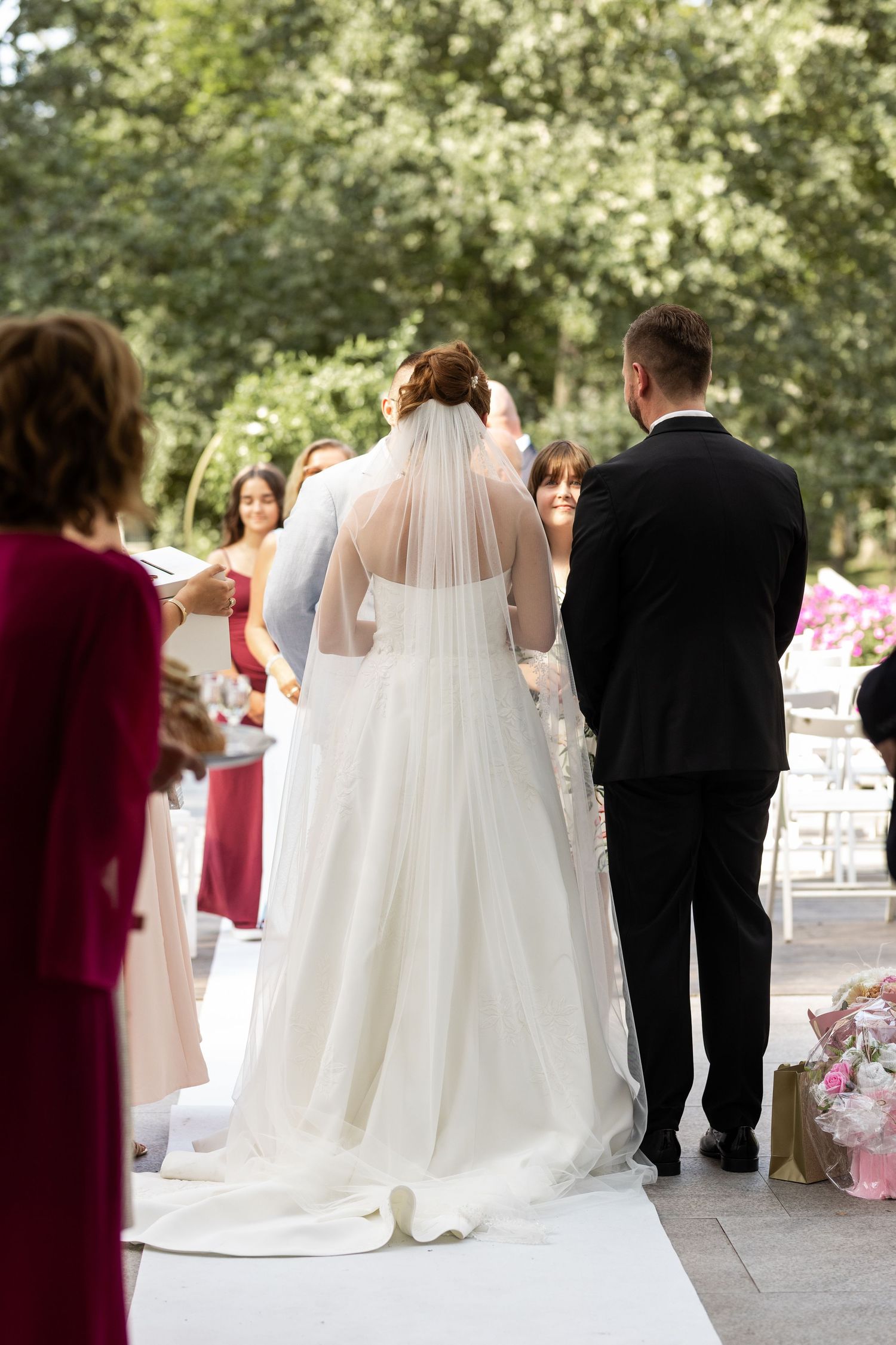 Couple stands at outdoor wedding ceremony with bride in flowing white gown and long veil on white aisle runner.
