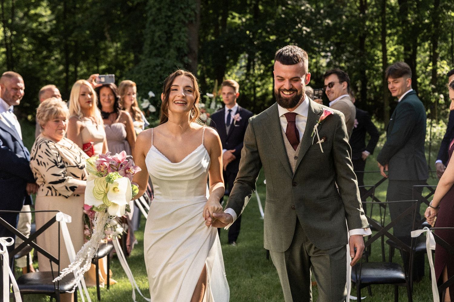 Wedding party laughing together while walking through a tree-lined outdoor path on sunny day.