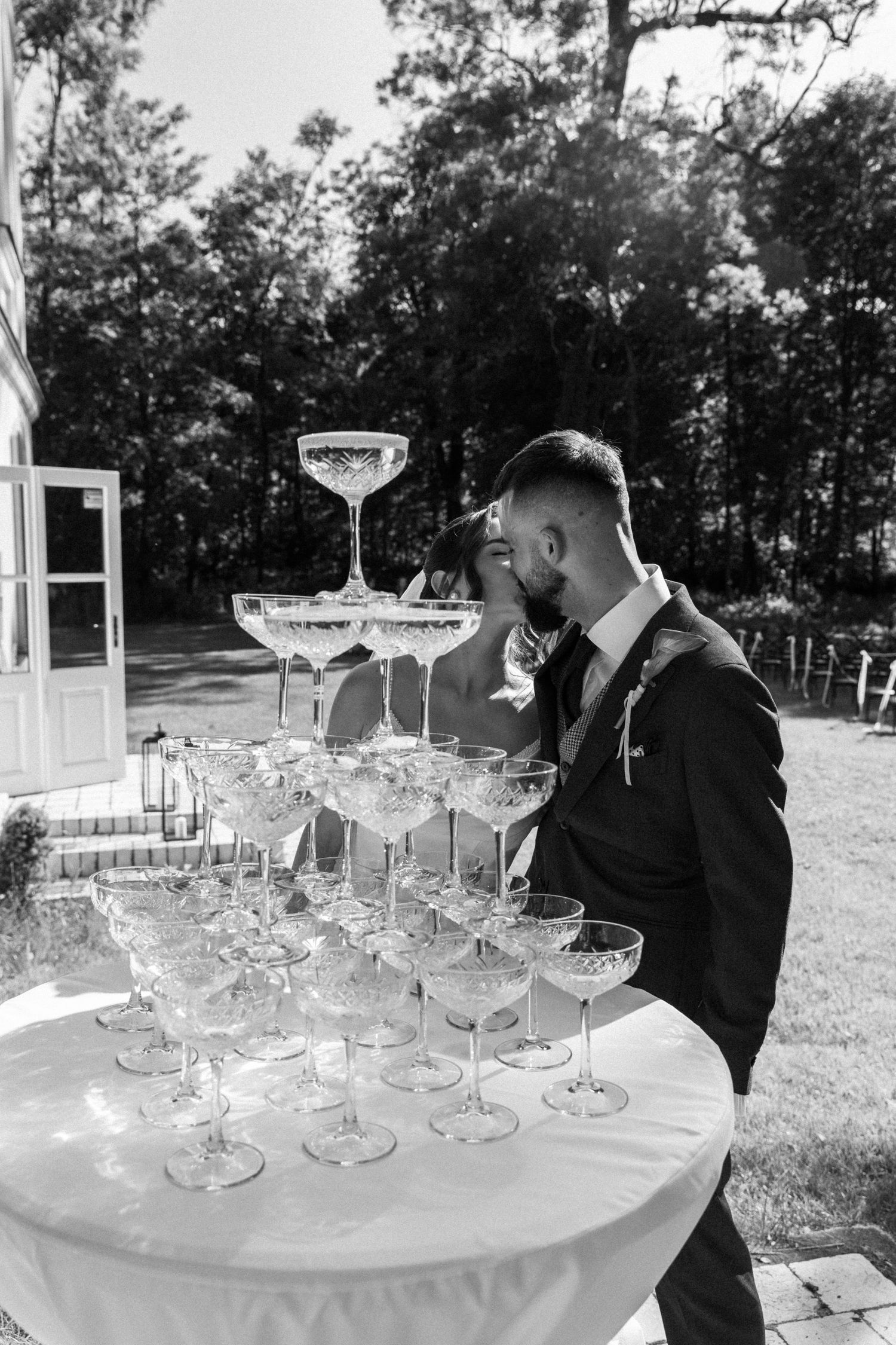 An elegant champagne tower setup at an outdoor wedding venue features stacked crystal coupe glasses ready to be filled.