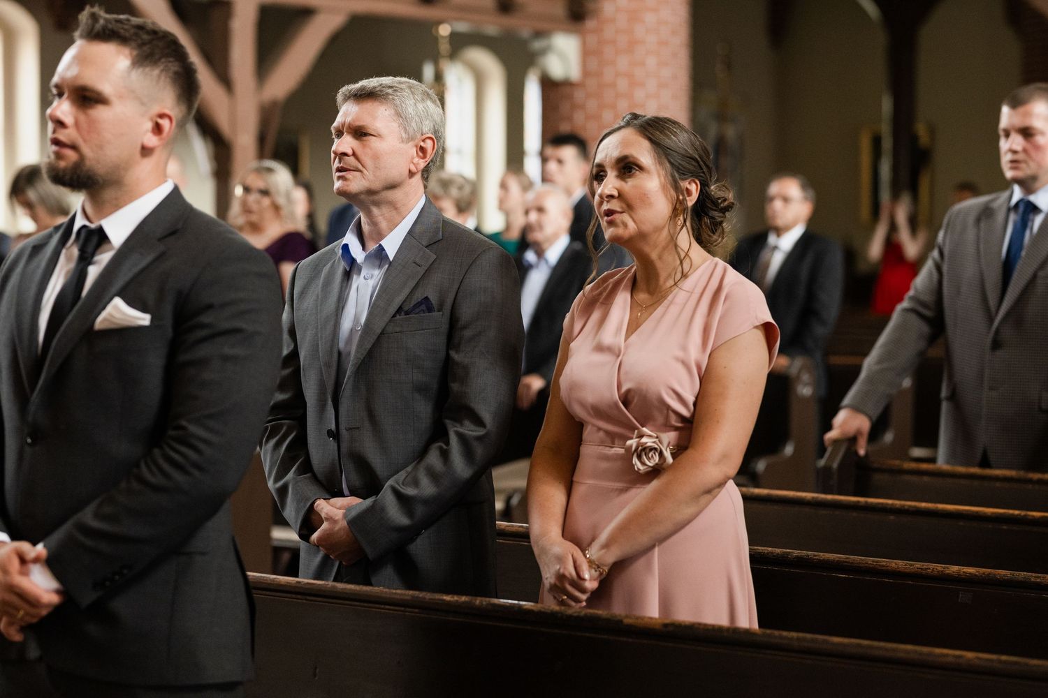 Wedding guests stand in church pews during a ceremony wearing formal attire in pink and dark suits.