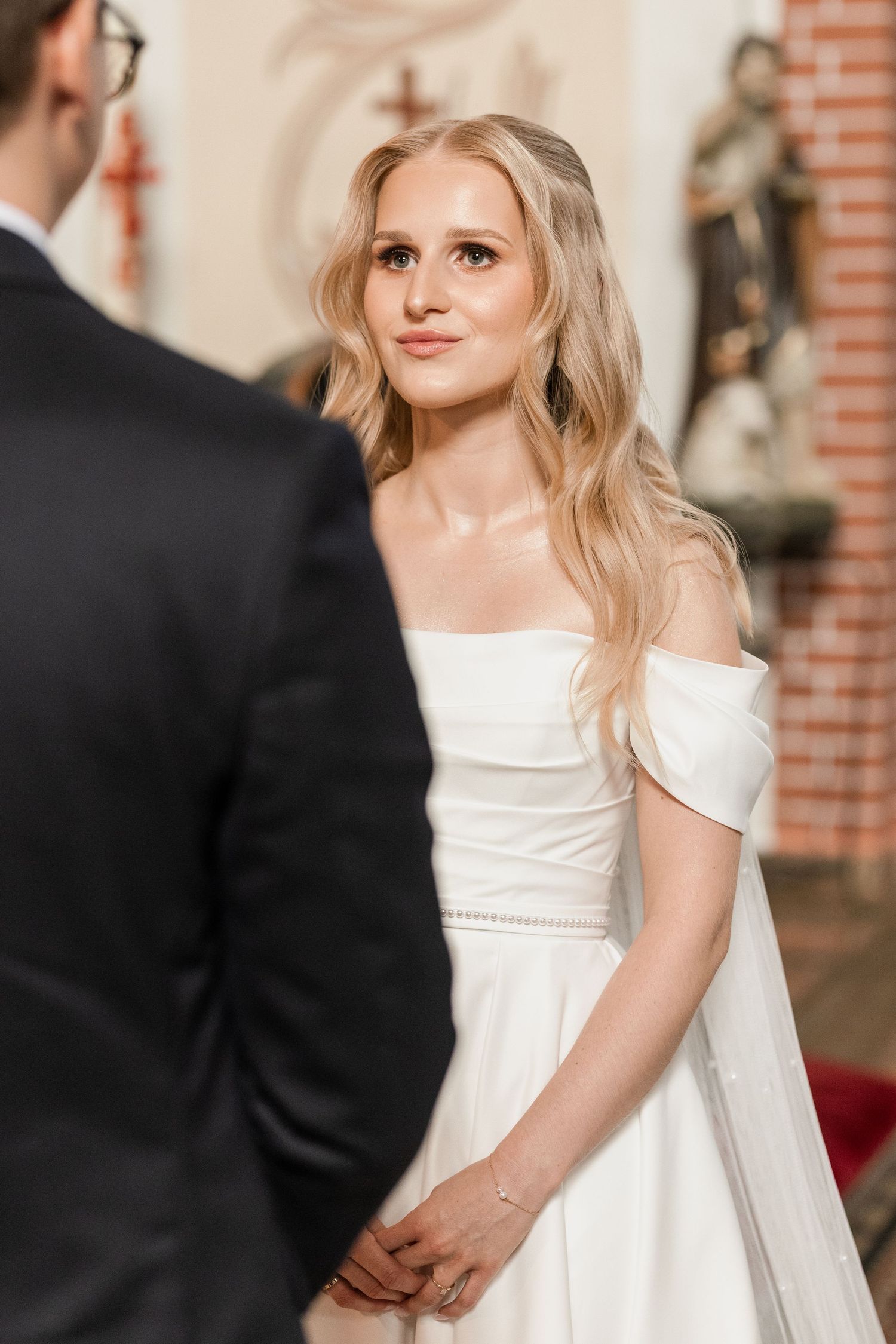 A bride in an elegant white wedding dress smiles while standing against a brick wall background.