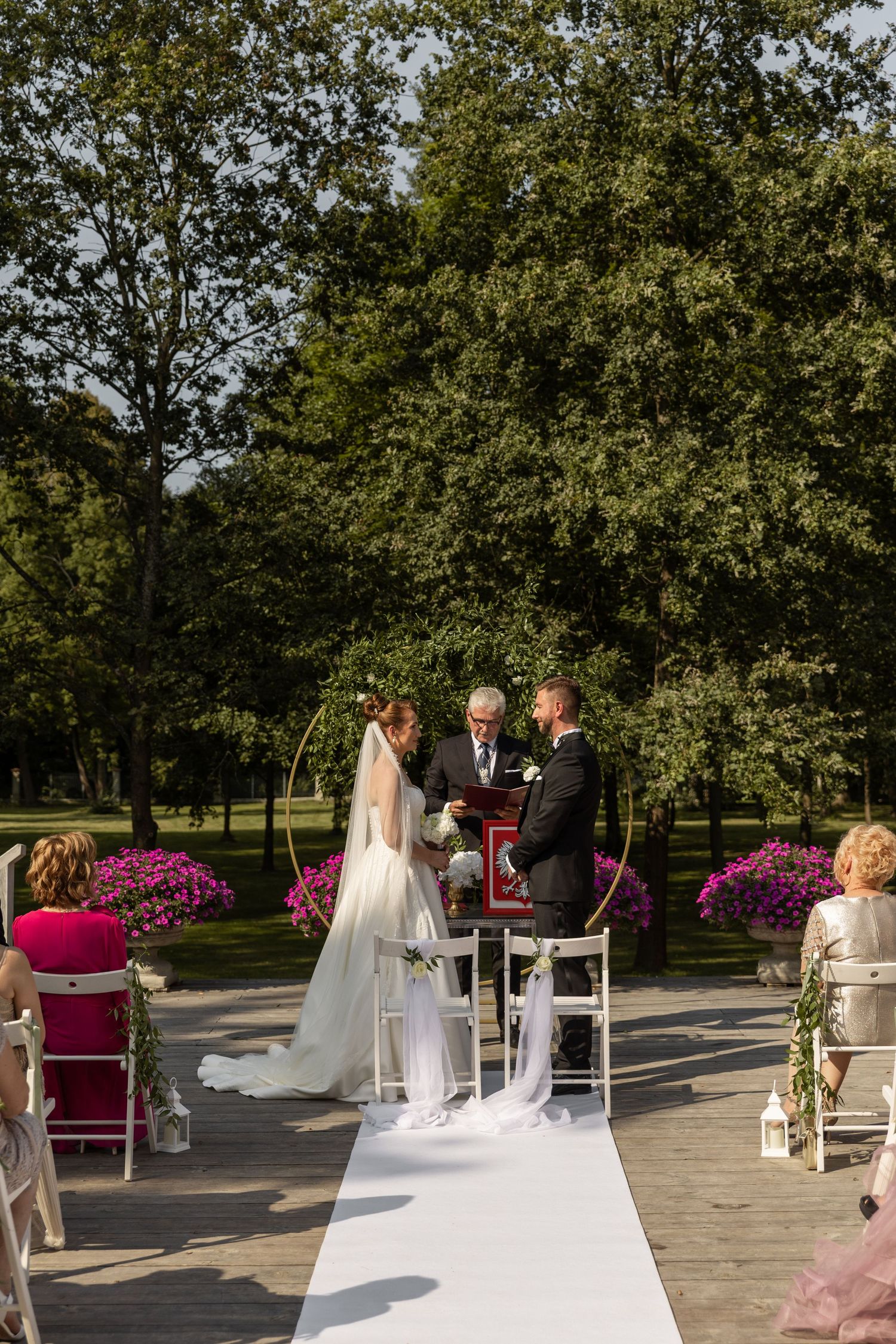 Outdoor wedding ceremony setup with white chairs, pink floral arrangements, and trees in the background.