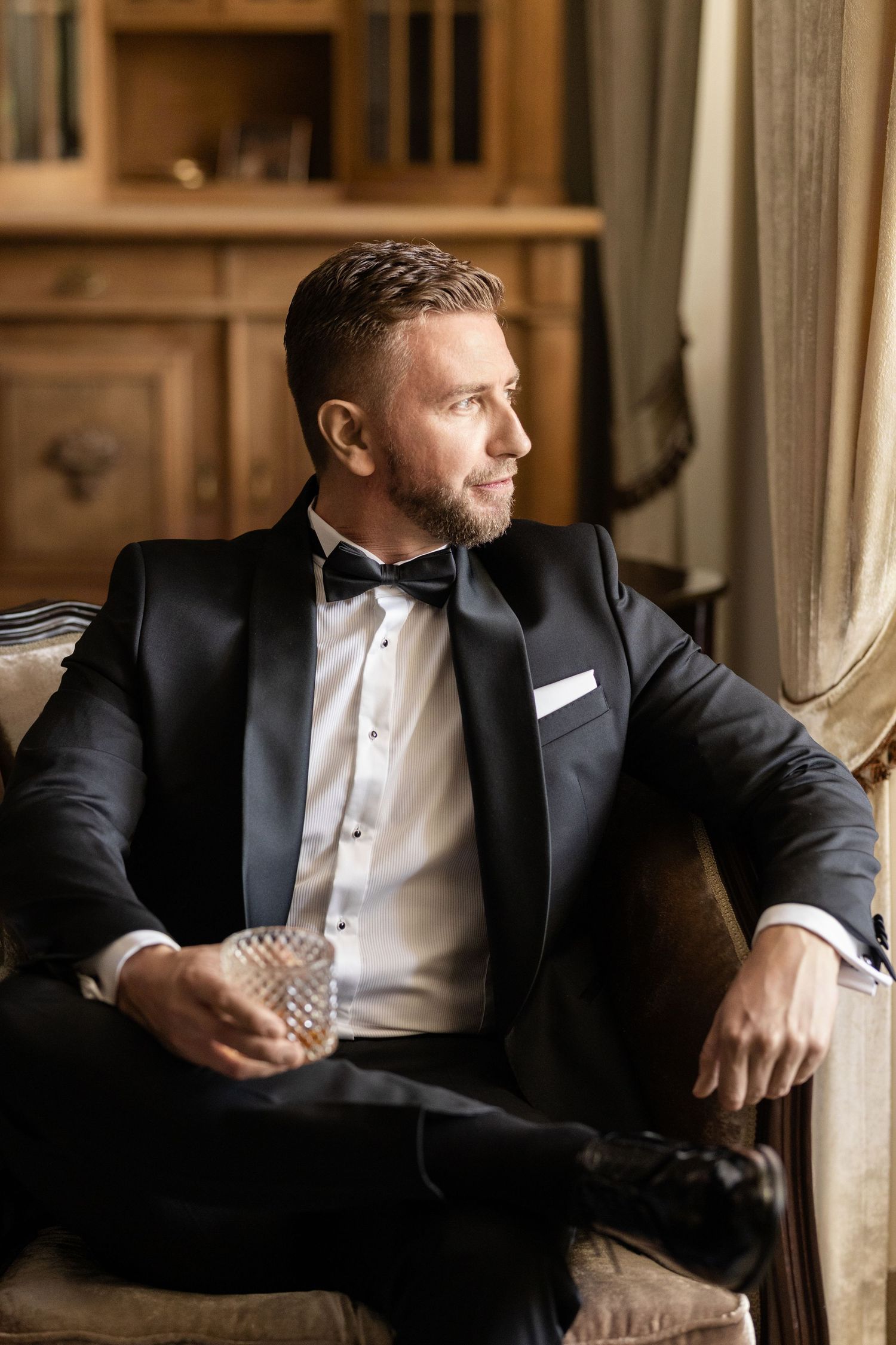 Man in black tuxedo sits by window in elegant setting gazing outward in contemplative pose.