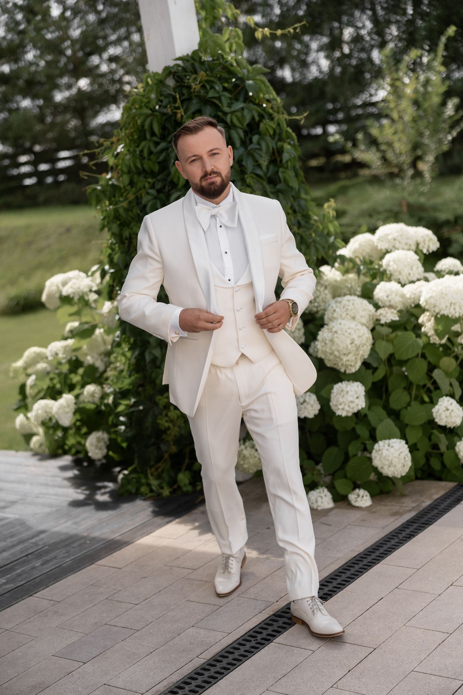 A groom in an ivory white suit poses confidently beside lush hydrangea bushes at an outdoor wedding ceremony.