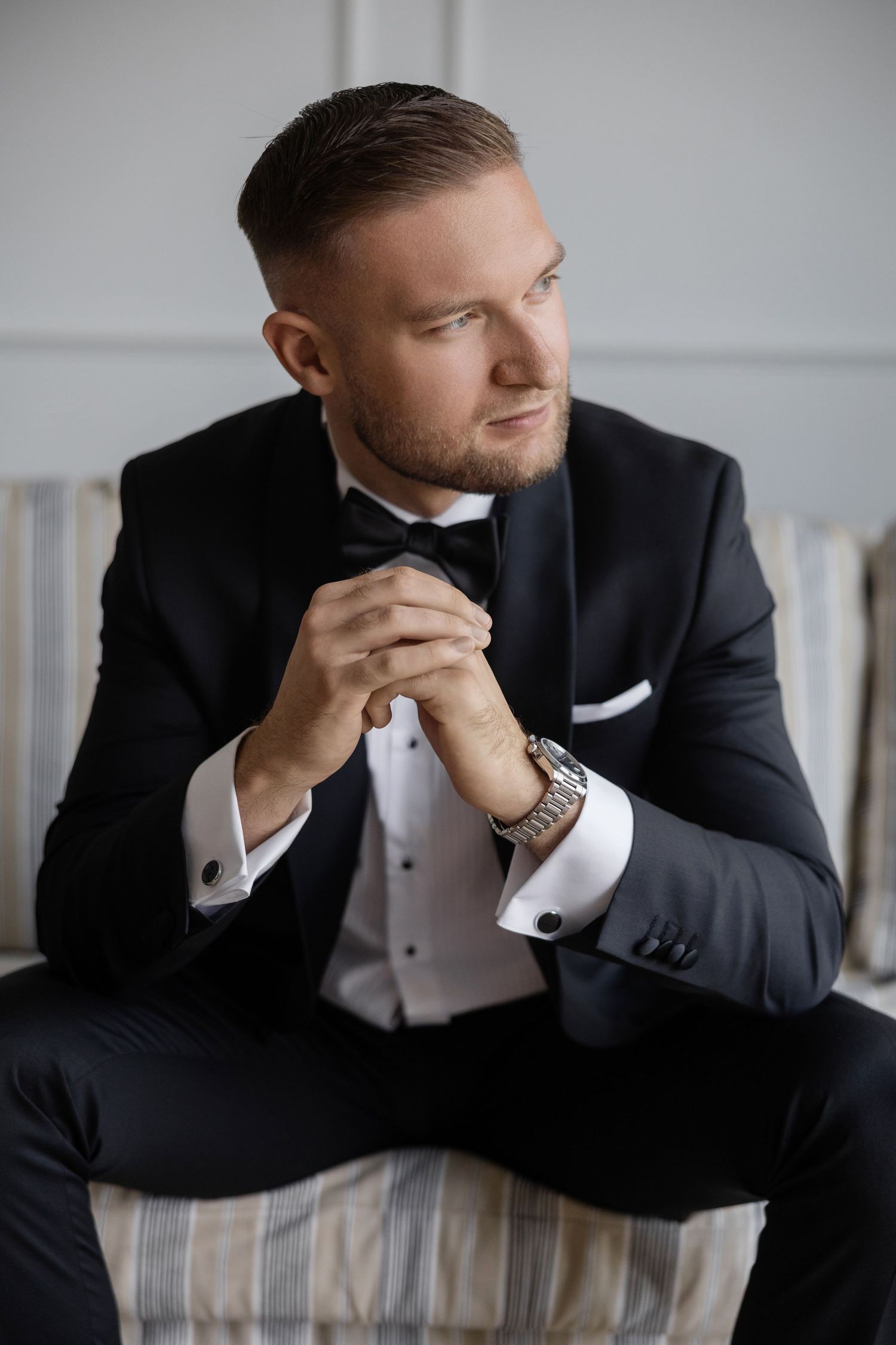 Elegant man in black tuxedo with bow tie and silver watch sits thoughtfully on striped sofa in formal attire.