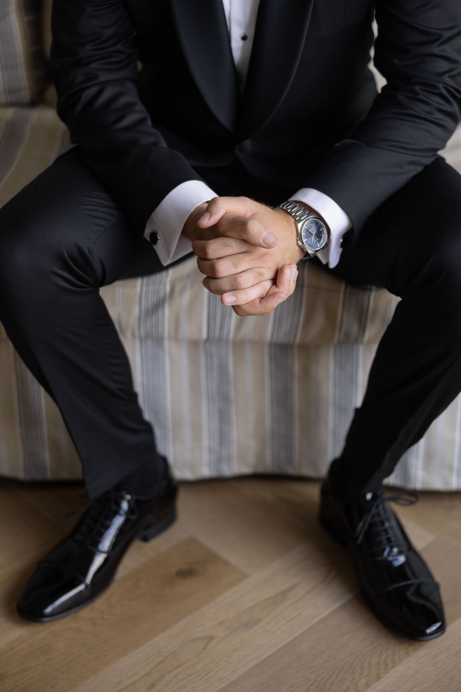 Man in black tuxedo with gold watch and cufflinks, seated with hands clasped, wearing patent leather oxford shoes.