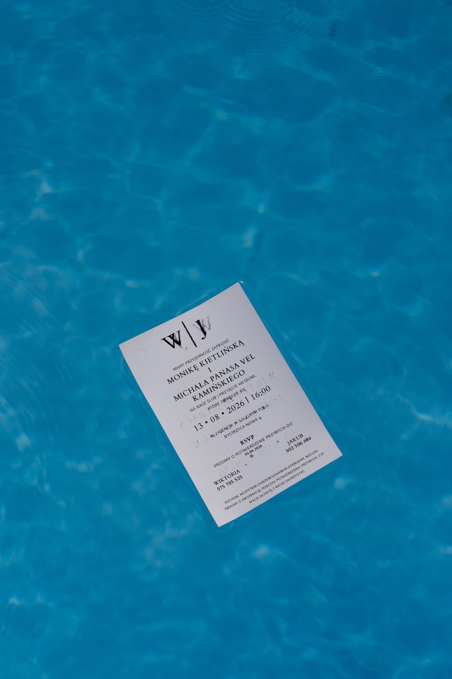 White wedding invitation card floating on bright blue pool water surface.