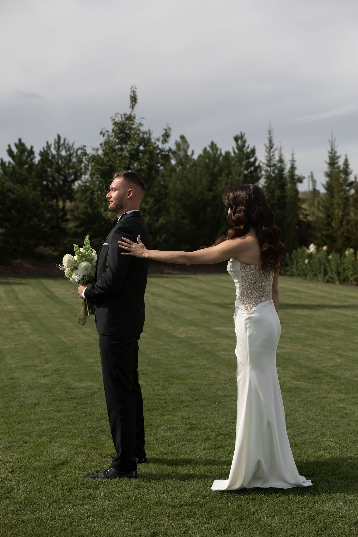 Bride in white gown touches groom's shoulder on green lawn with evergreen trees backdrop.