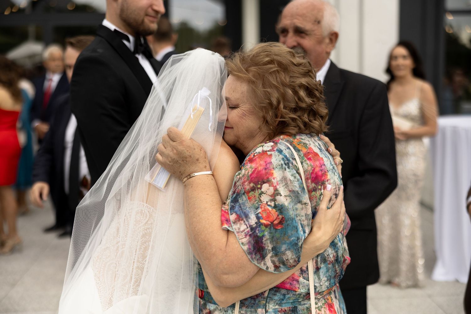 Bride in white gown and veil sharing an emotional hug with older woman in floral dress at outdoor wedding reception.