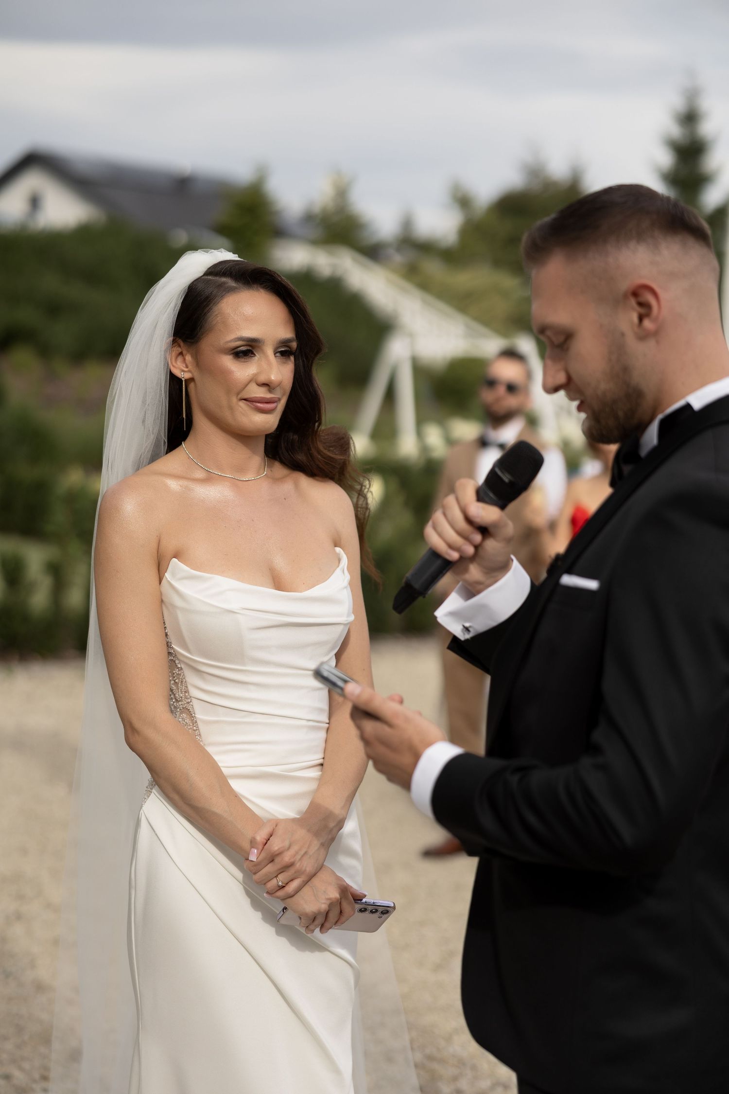 Groom reads wedding vows to emotional bride in white strapless gown during outdoor ceremony.