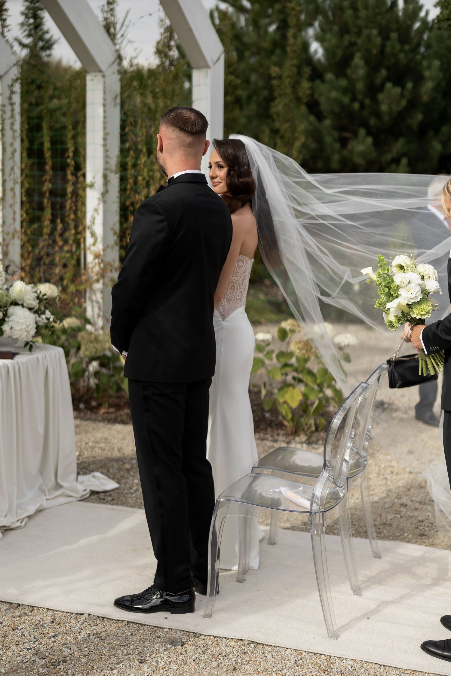 Bride in white gown and veil holds bouquet facing groom in black tuxedo at outdoor wedding ceremony.