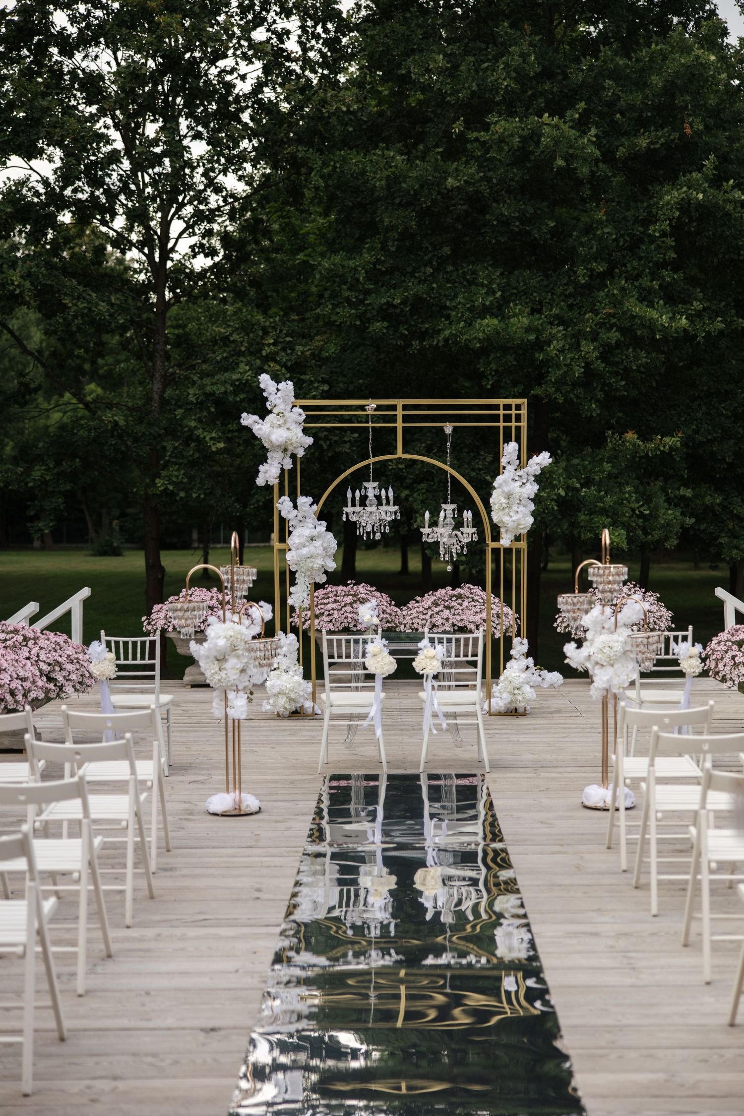 Elegant outdoor wedding ceremony setup with gold arch, white chairs, chandelier, and floral arrangements on a wooden deck.