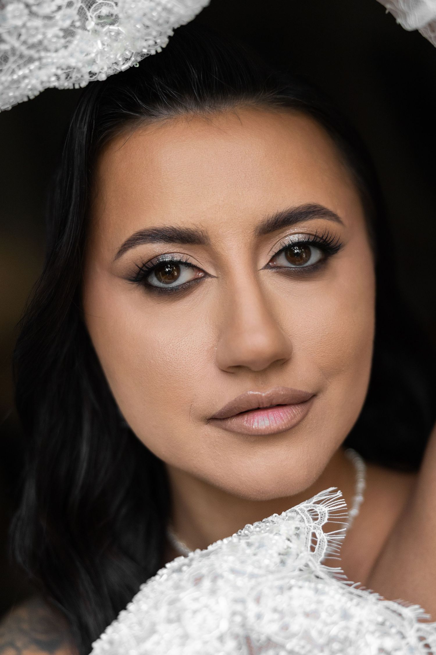 Close-up portrait of a bride with dark hair, smoky eye makeup, and white lace wedding dress and floral headpiece.