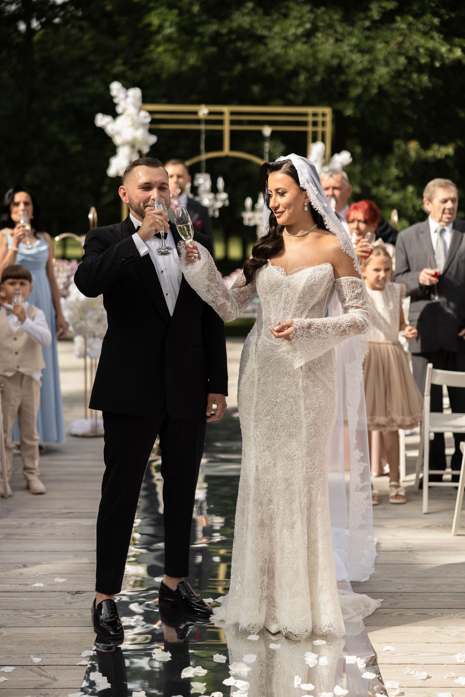 Bride and groom toasting with champagne glasses at outdoor wedding ceremony, guests seated behind them.