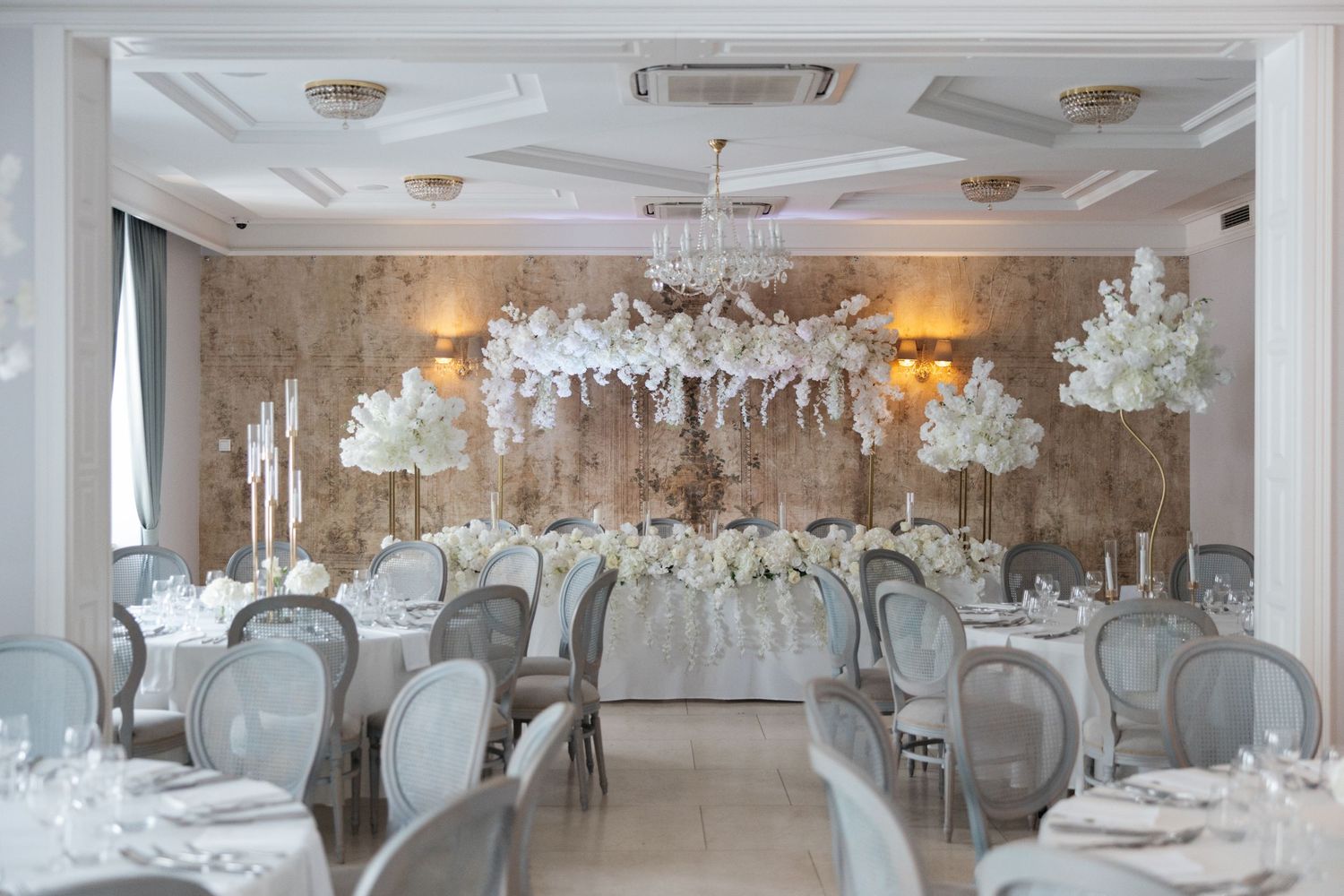 Elegant wedding reception hall with white floral centerpieces, gray chairs, chandelier, and ornate wall decor.