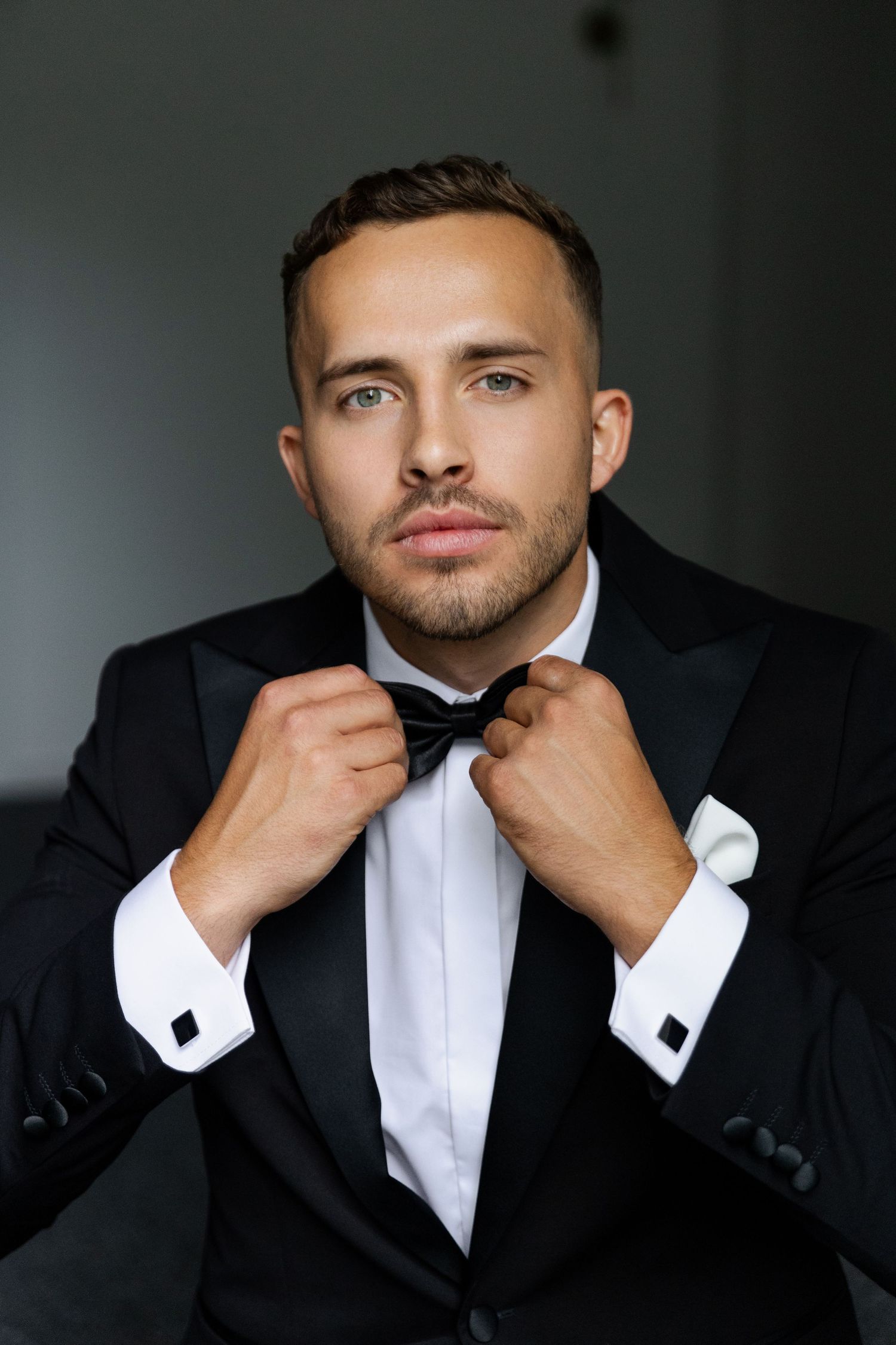 Professional portrait of a man adjusting his bowtie while wearing a classic black tuxedo and white dress shirt.