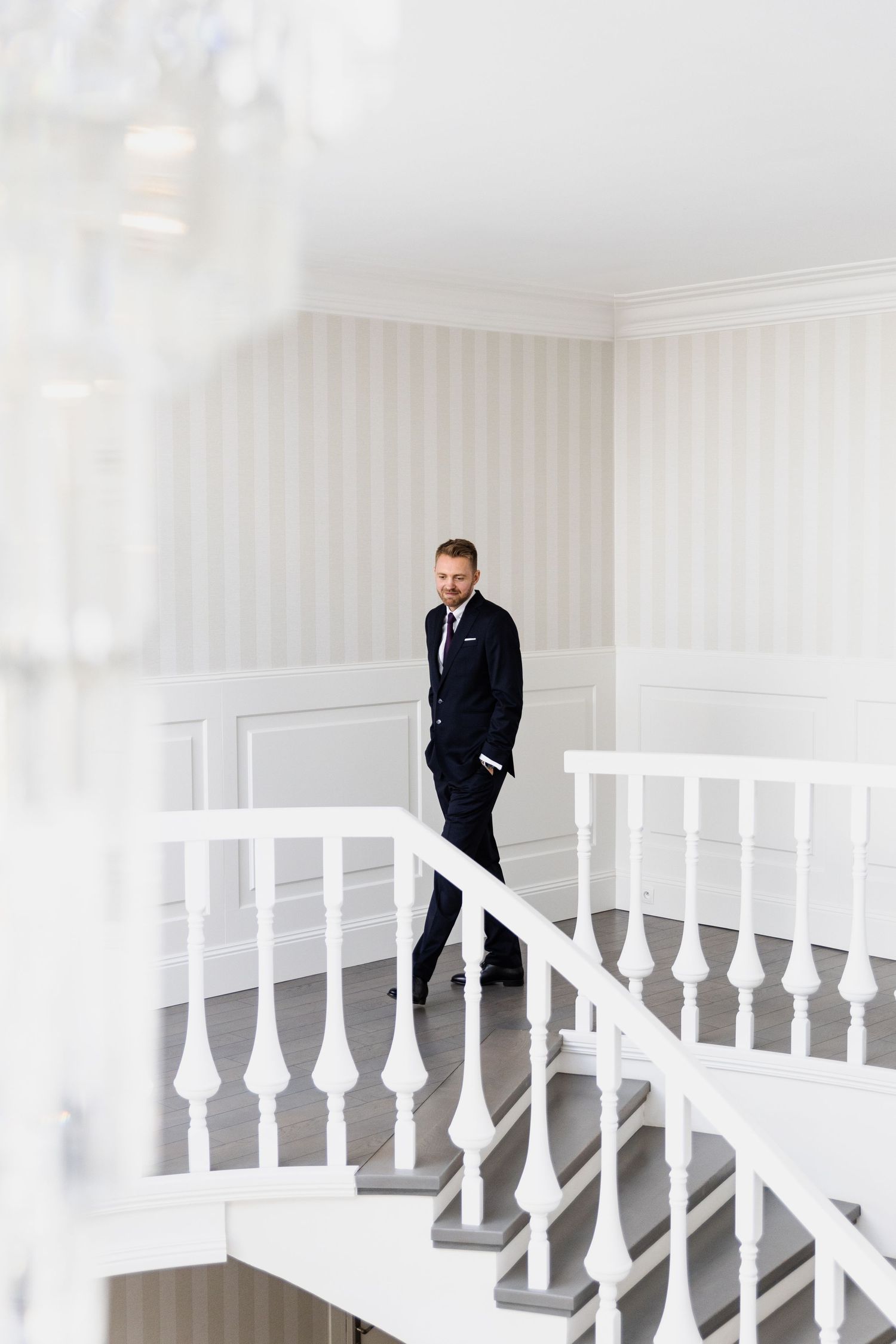 A person in a black suit stands on an elegant white staircase with ornate balusters in a bright interior space.