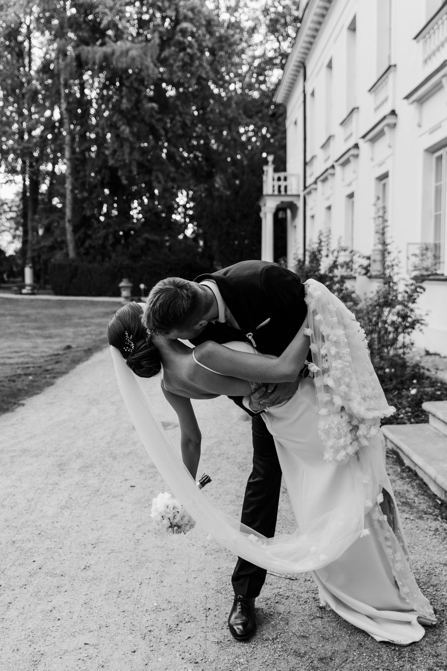 A romantic black and white photo showing a couple in wedding attire sharing an intimate dip pose outside a grand building.