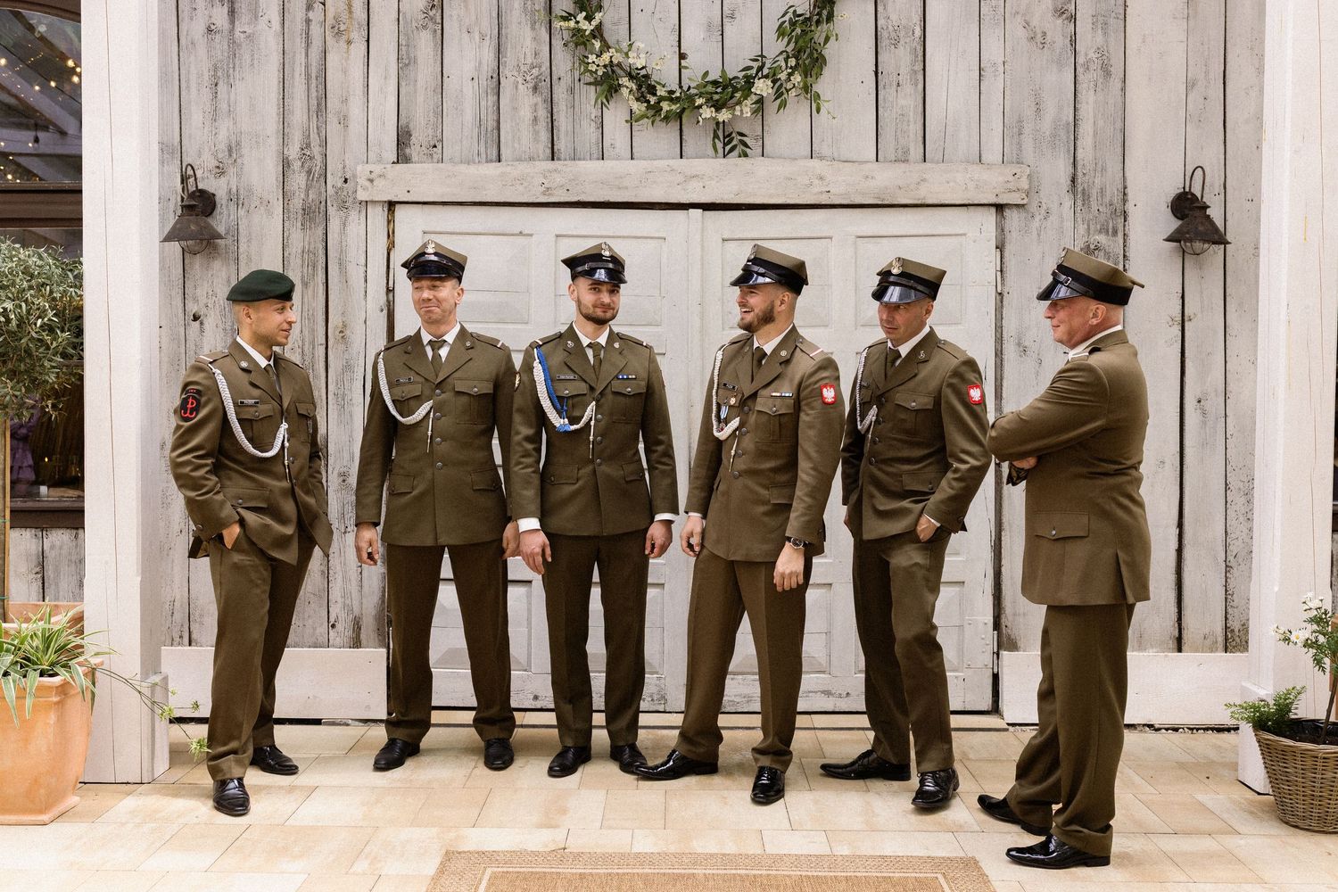 Groomsmen wearing olive military dress uniforms stand together in front of rustic white barn door with greenery.