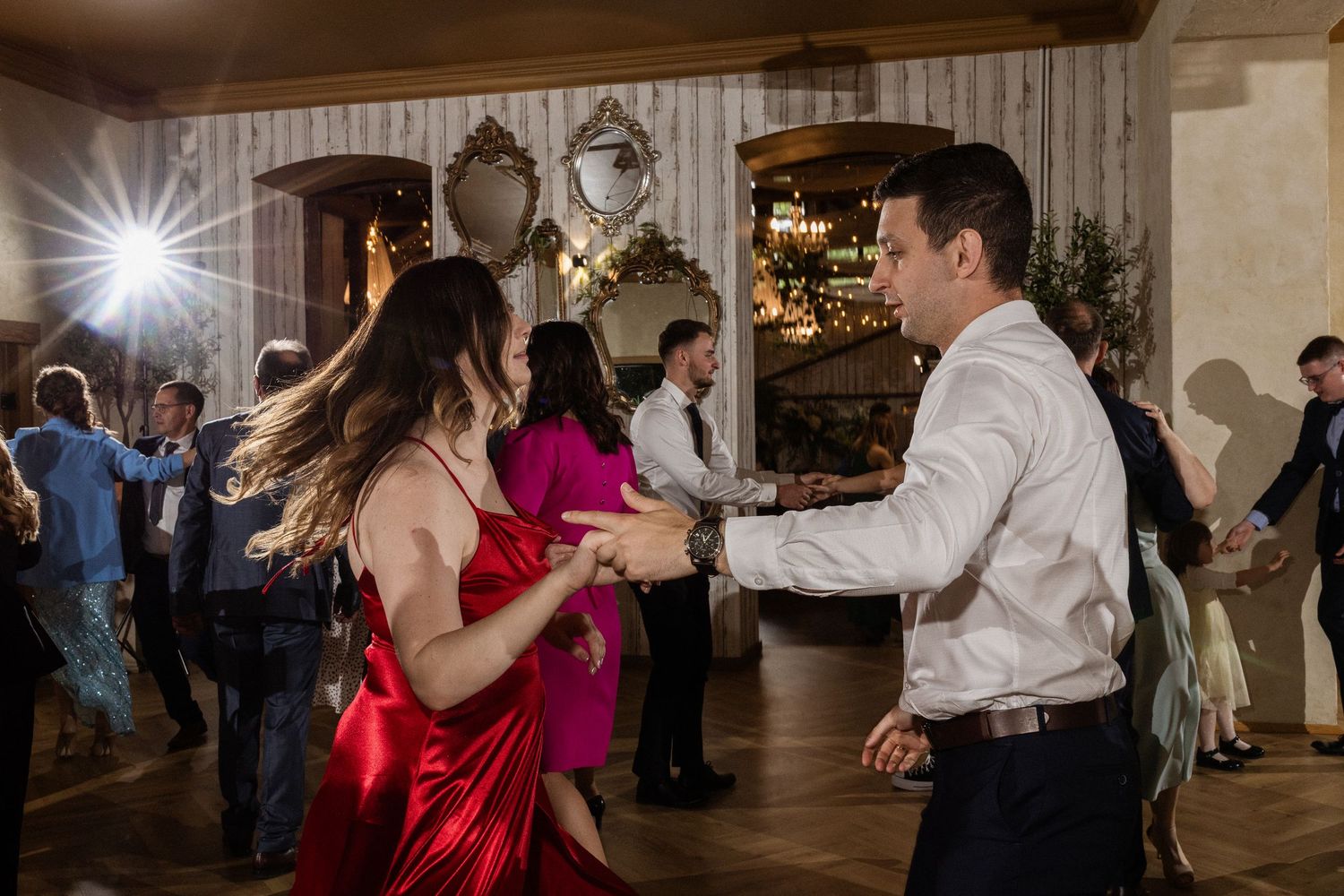 Guests dancing enthusiastically at a wedding reception venue with decorative lights and mirrors on the wall.